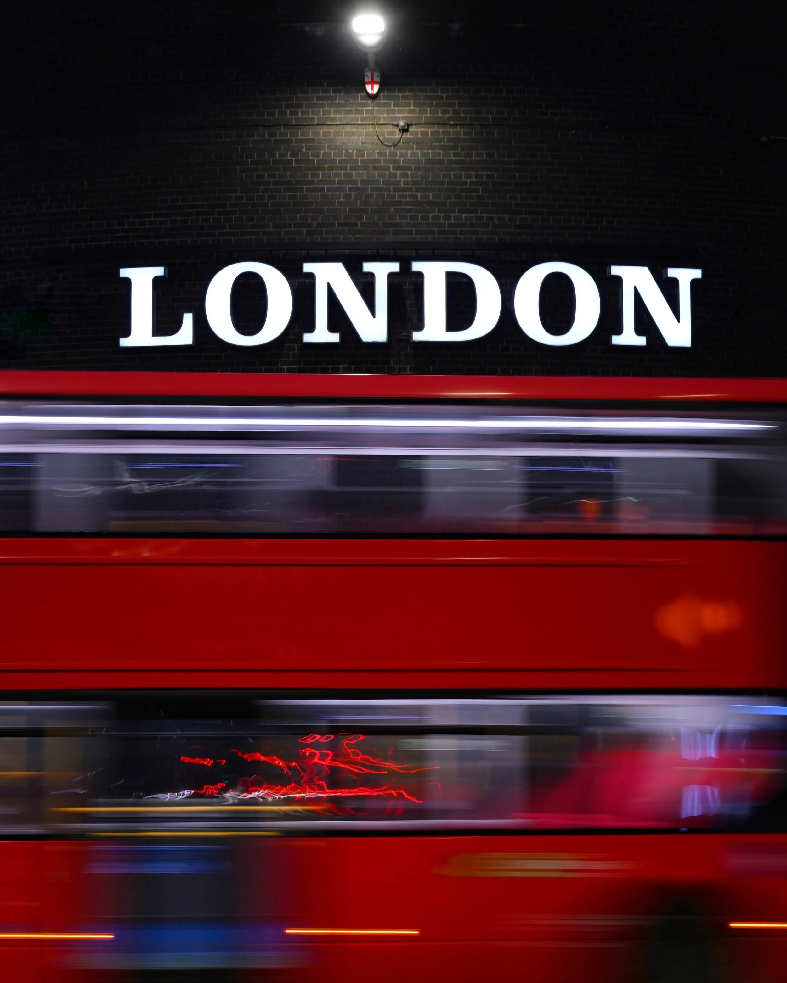 Une photo de nuit de l'enseigne lumineuse 'LONDON' sur un mur en briques, avec un éclairage d'une lampe au-dessus. La photo montre également un mouvement flou des voitures passant devant.