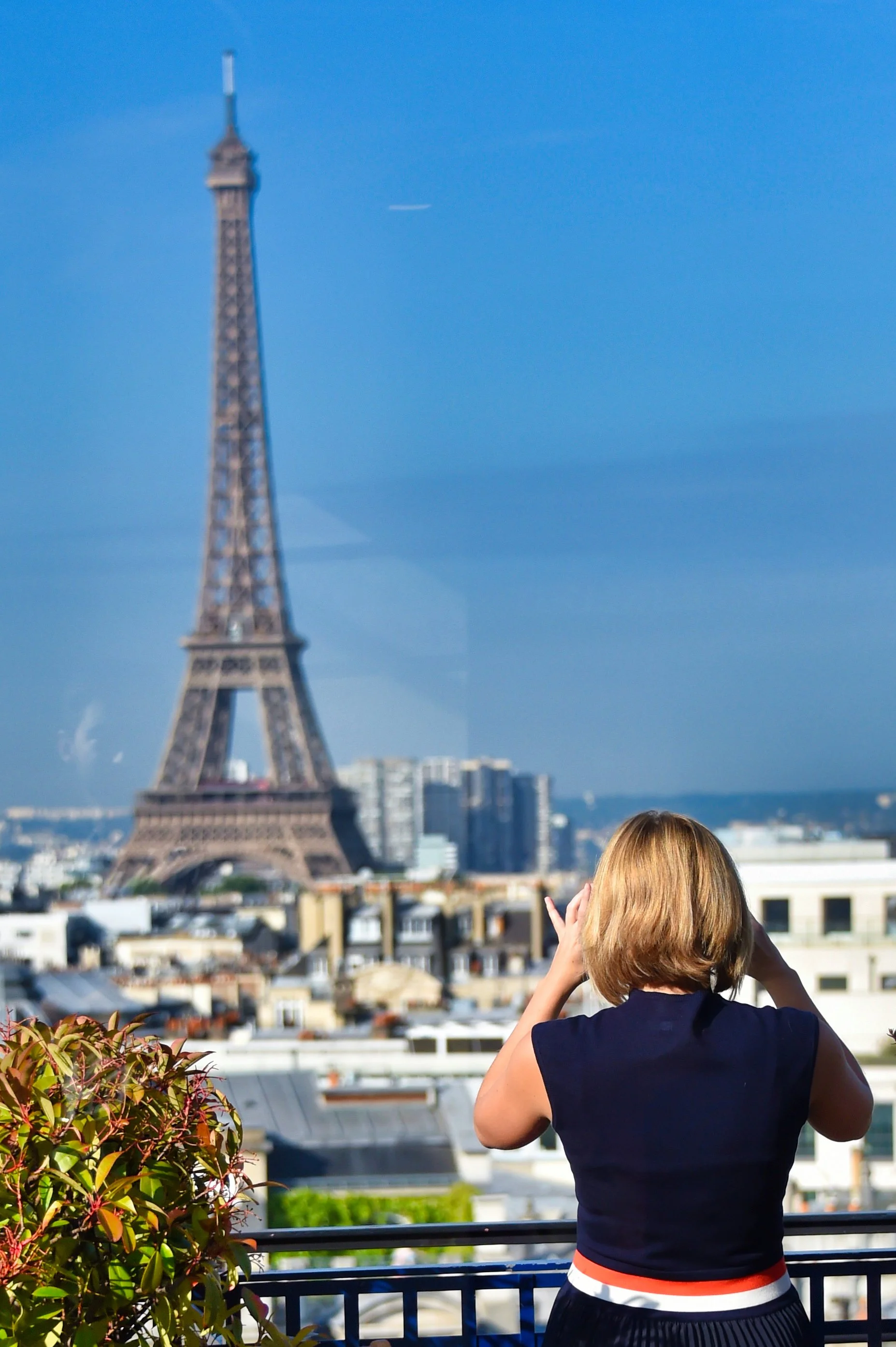 Personne avec cheveux courts, portant un haut noir avec une bande blanche rouge, prenant une photo avec un smartphone devant la tour Eiffel à Paris, en journée avec un ciel bleu.