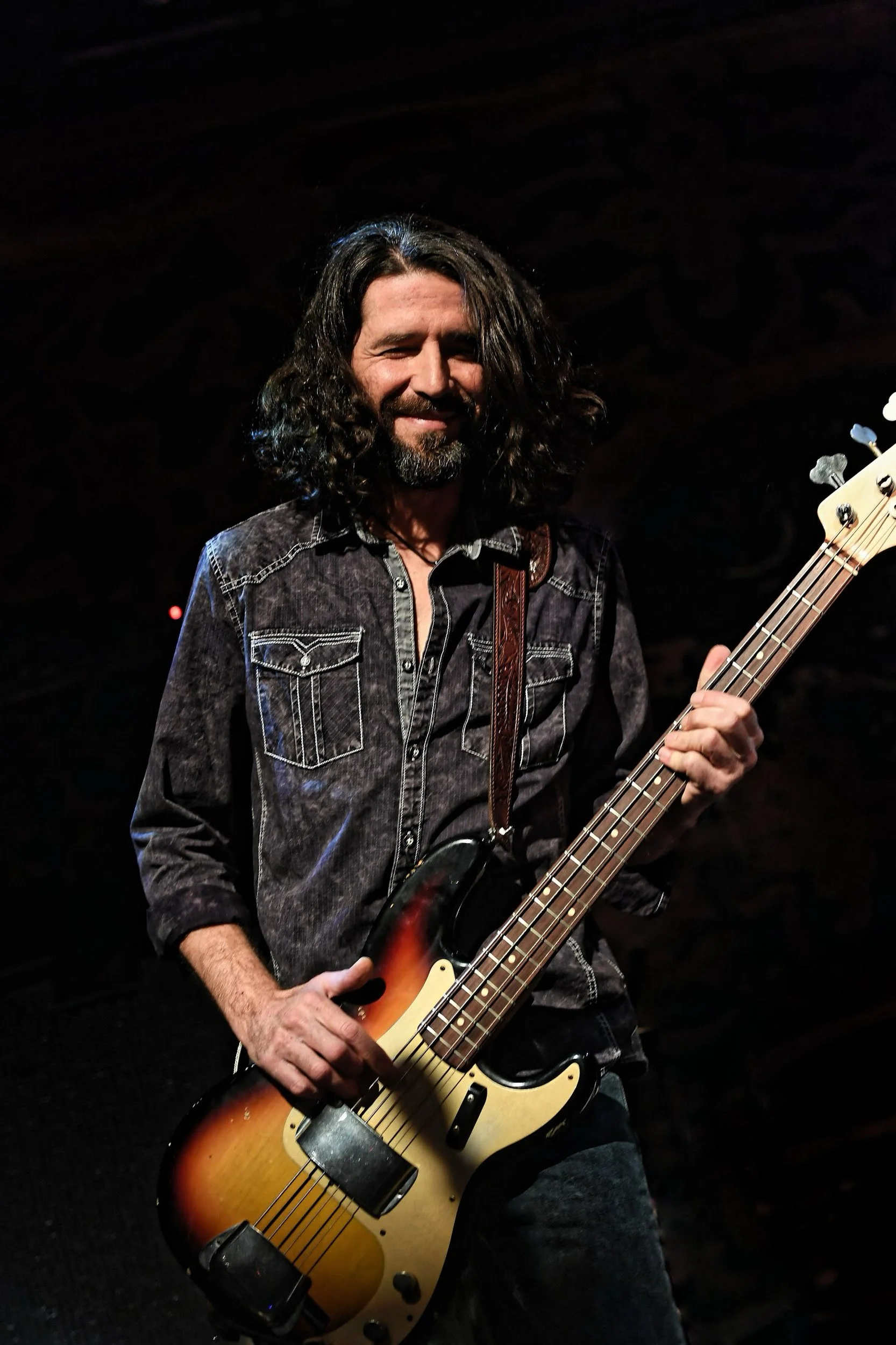 Un homme avec des cheveux longs et une barbe joue de la basse électrique sur scène, portant une veste en denim noire, dans un environnement sombre.
