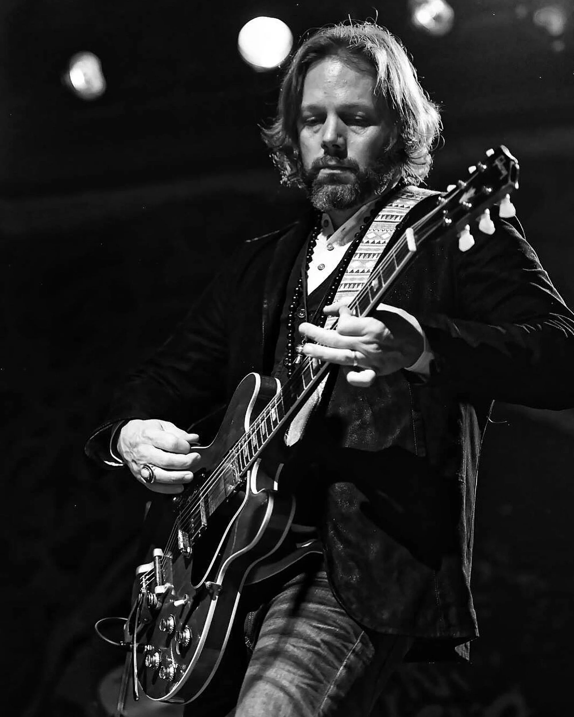 Un homme avec des cheveux longs et une barbe joue de la guitare électrique lors d'une performance, en noir et blanc, avec un arrière-plan flou.