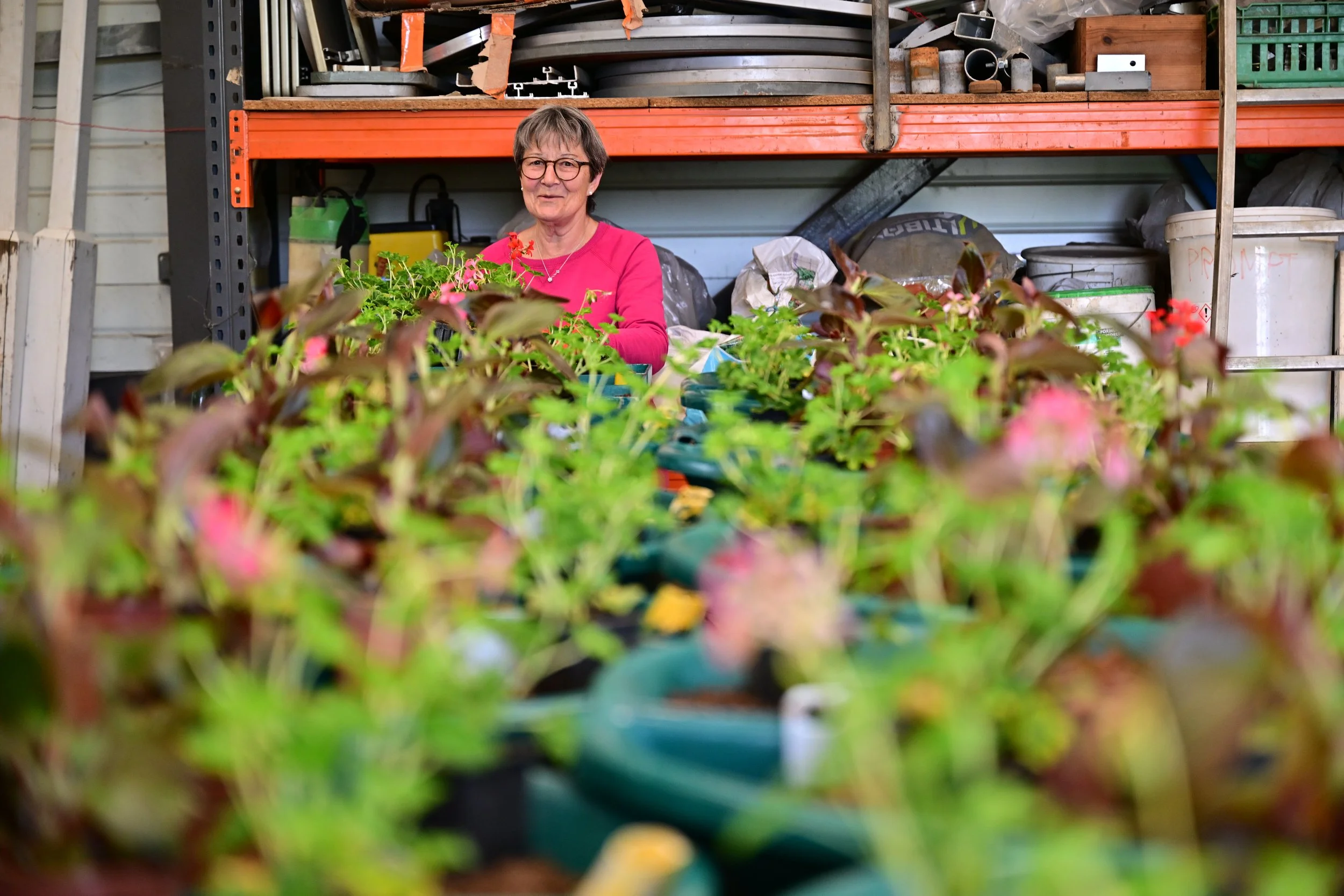 Une femme souriante portant des lunettes, travaillant parmi des plantes en pot dans une serre ou un atelier de jardinage, avec des étagères contenant divers outils et matériaux en arrière-plan.