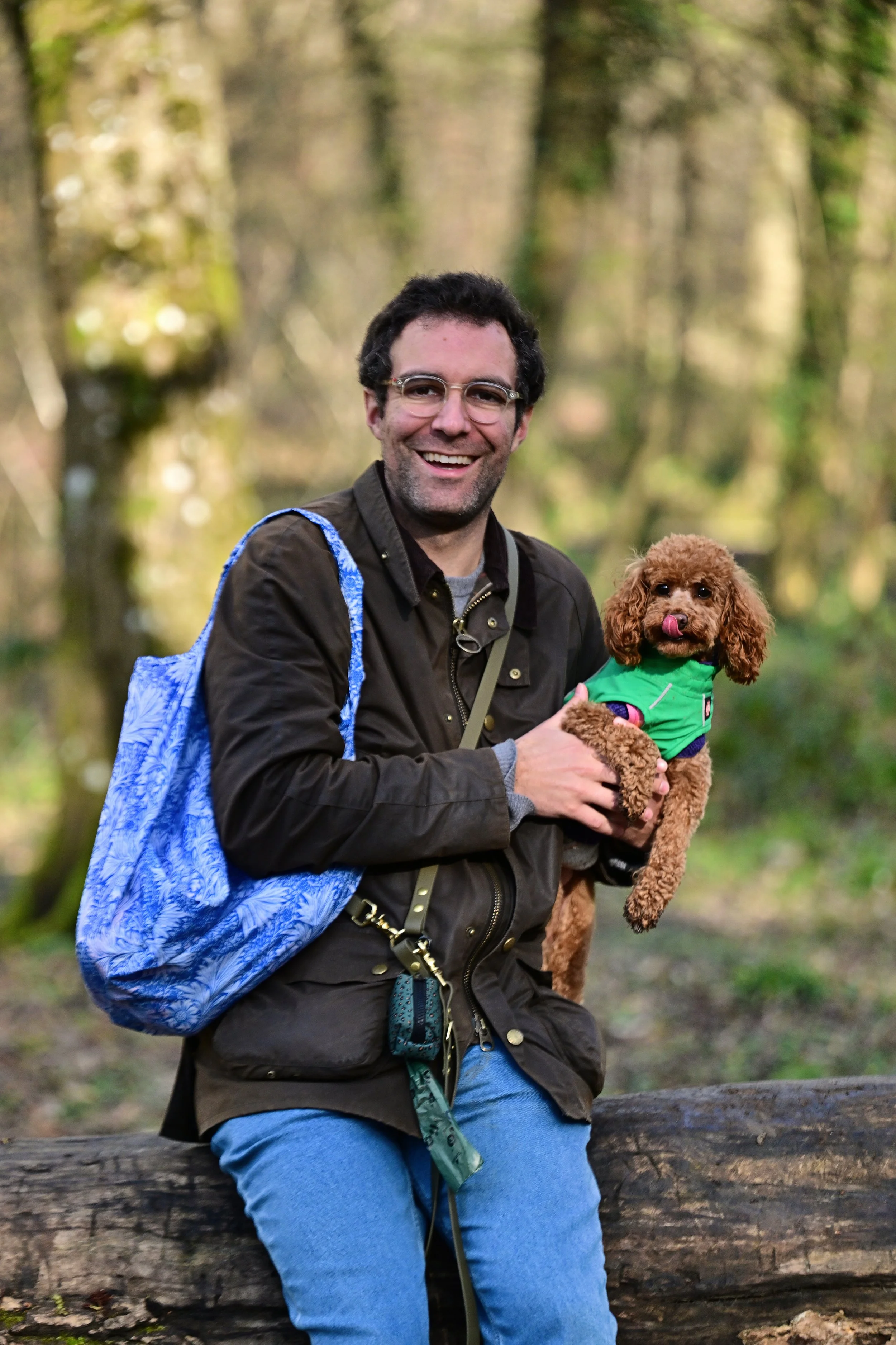 Un homme souriant, portant des lunettes, avec un sac à dos bleu, tient un petit chien pékinois marron portant un gilet vert dans un parc forestier avec des arbres en arrière-plan.