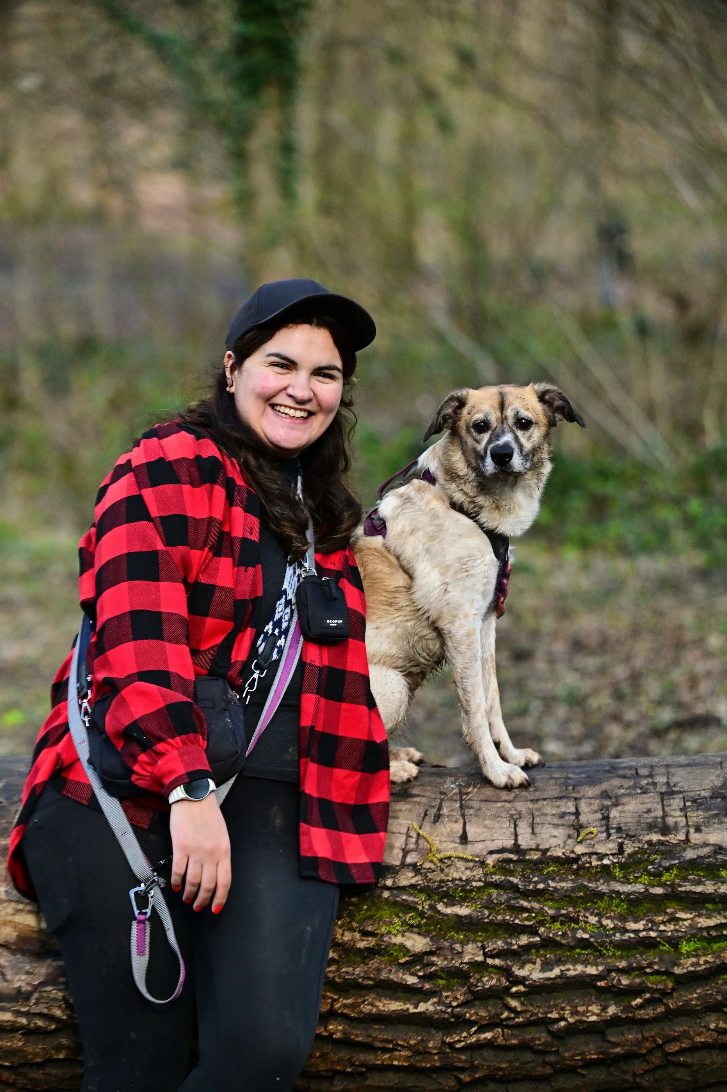 Une femme souriante avec un casque noir porte une veste à carreaux rouges et noirs, tenant un chien debout sur une grande bûche en bois en forêt.
