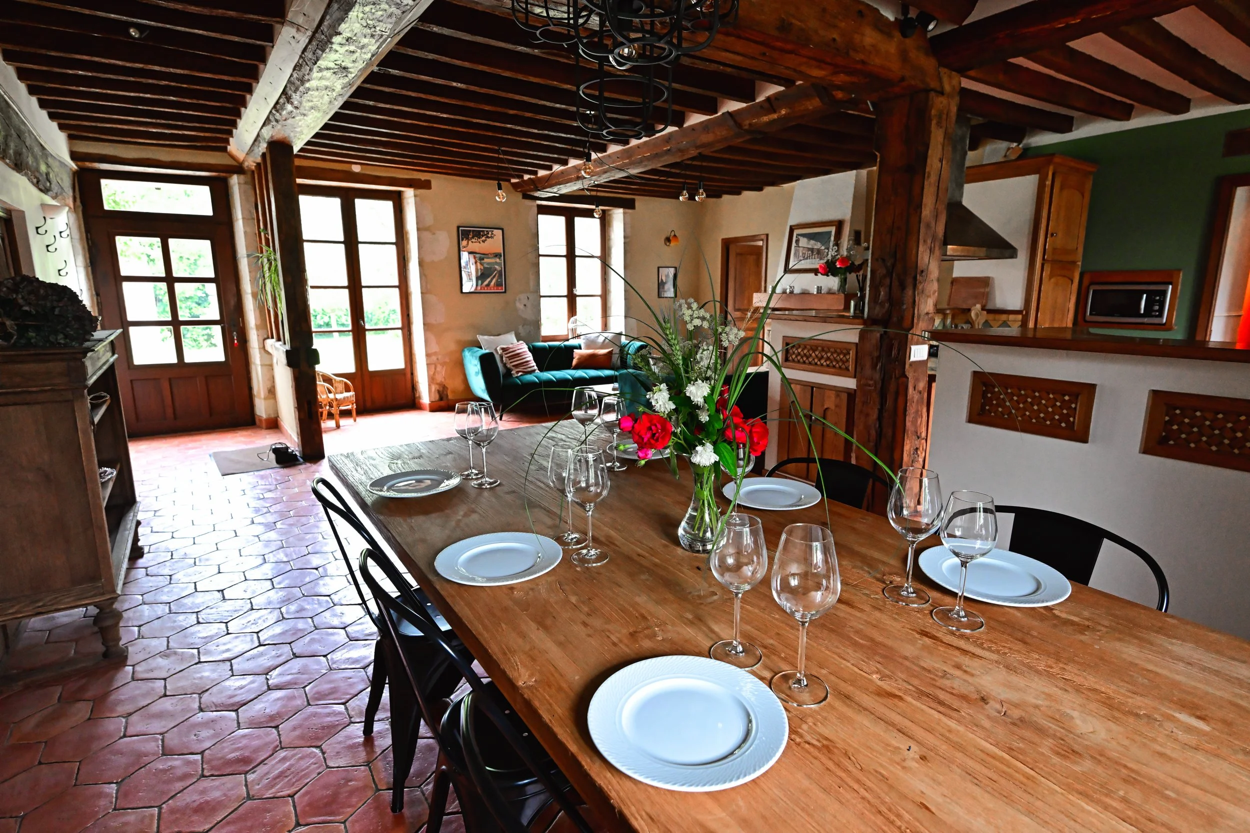 Une salle à manger chaleureuse avec un long tapis en terre cuite au sol, un grand tableau, des fenêtres en bois laissant entrer la lumière naturelle, une table en bois avec des verres et des assiettes, et un vase avec des fleurs colorées au centre.