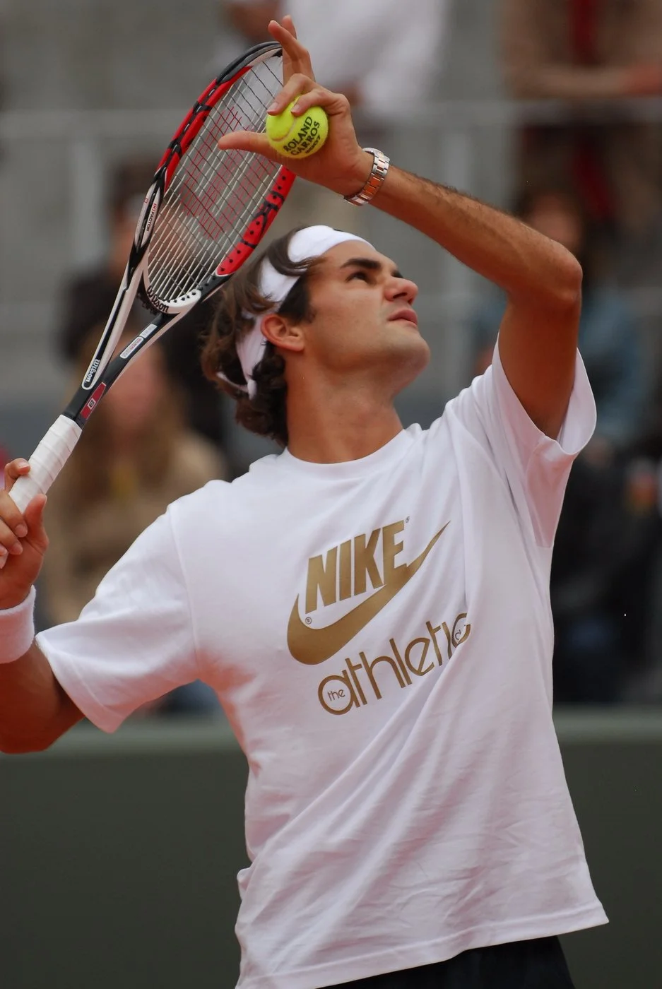 Un homme joue au tennis, portant un t-shirt blanc Nike avec le logo or et le mot 'theathletic' en dessous, une raquette de tennis en main et une balle de tennis jaune dans l'autre main, lors d'un match ou entraînement.