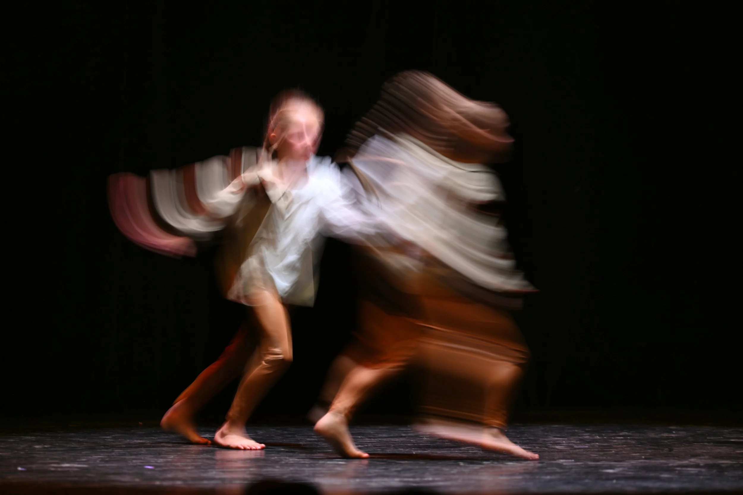 Deux danseurs en mouvement sur scène, vêtus de costumes amples, flous à cause du mouvement, fond noir.