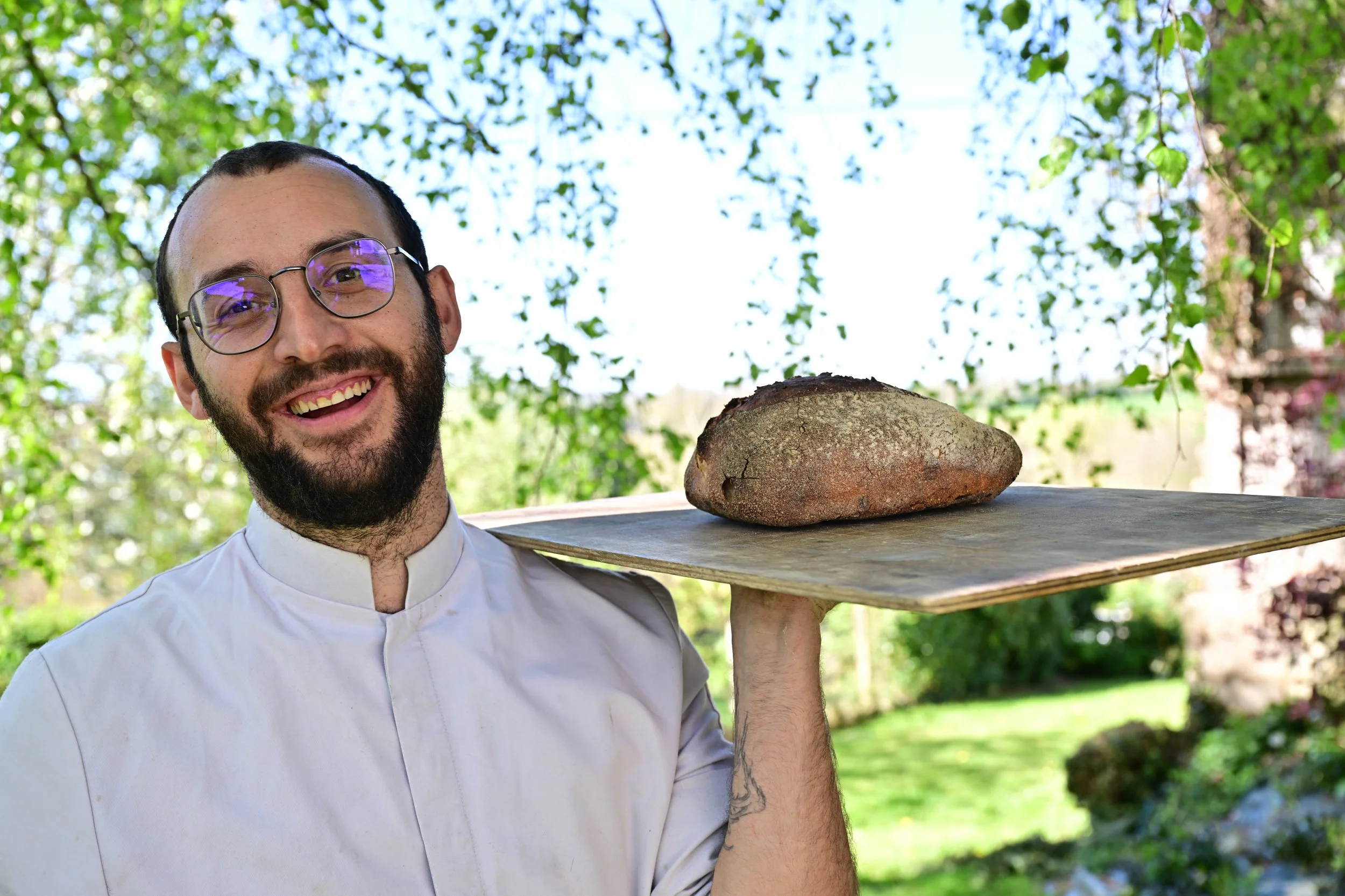 Un homme souriant avec des lunettes porte un plateau avec une grande pierre lors d'une activité en plein air, avec un fond de feuilles vertes.