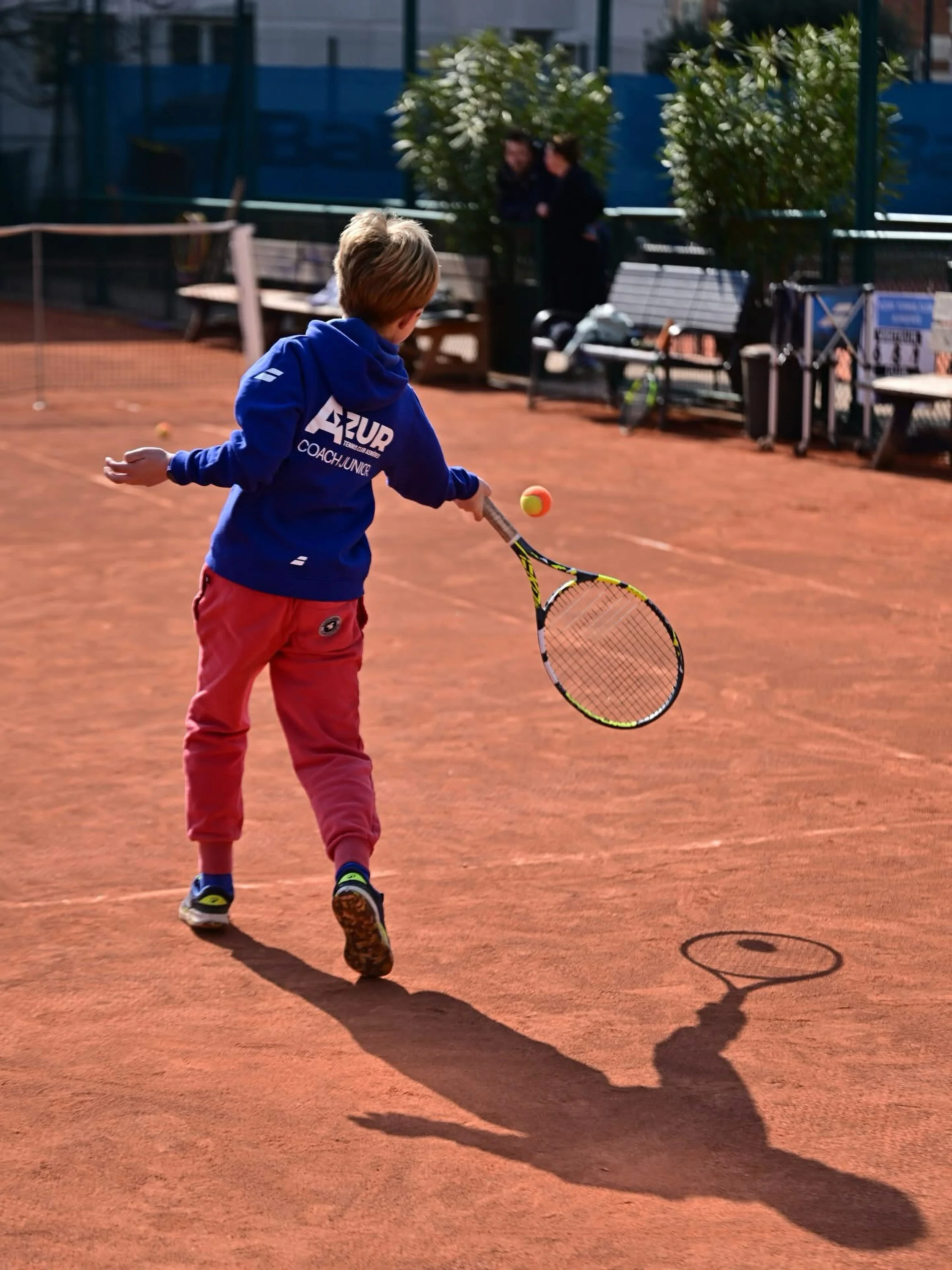Un jeune garçon joue au tennis sur un court en terre battue, avec deux femmes assises en arrière-plan et un arbre, en plein jour.