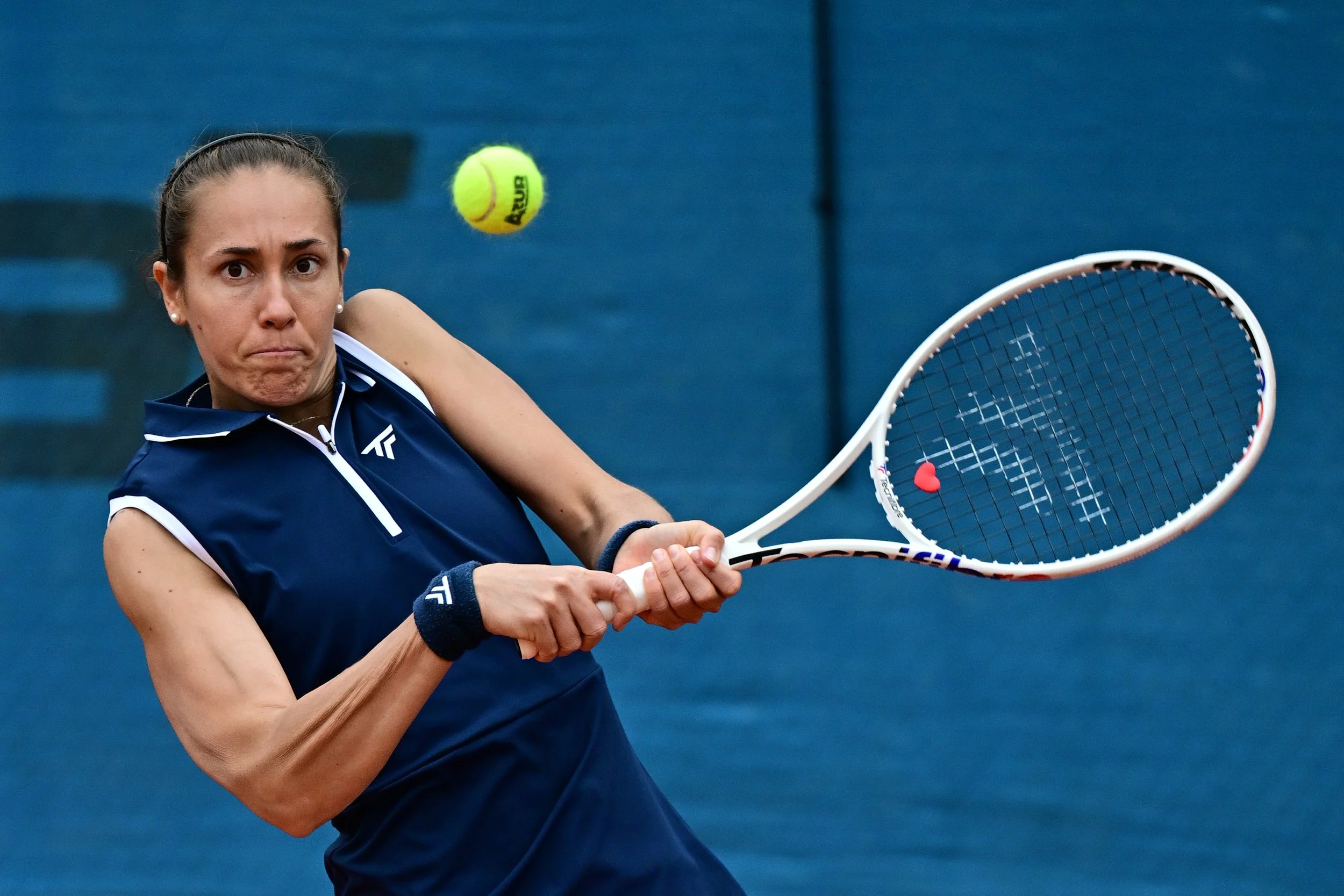 Une femme joue au tennis, prête à frapper une balle jaune en plein mouvement, avec un fond bleu.
