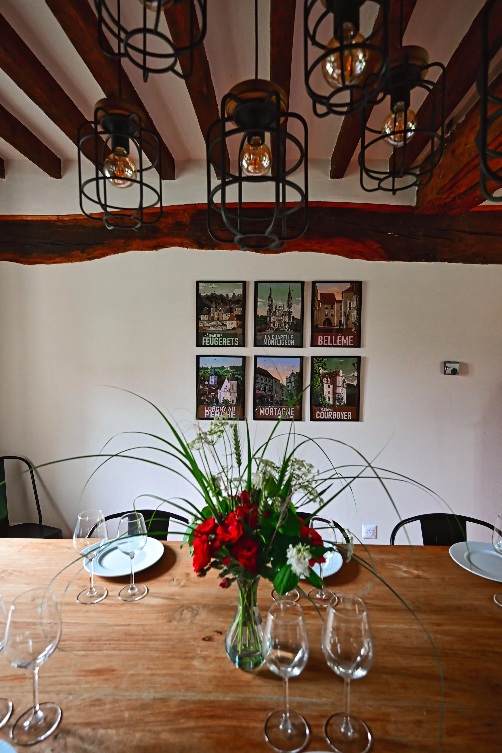 Une table en bois avec des verres à vin et des assiettes. Au centre, un bouquet de fleurs rouges et blanches avec des feuille vertes dans un vase en verre. En arrière-plan, des affiches de villes françaises sur le mur.