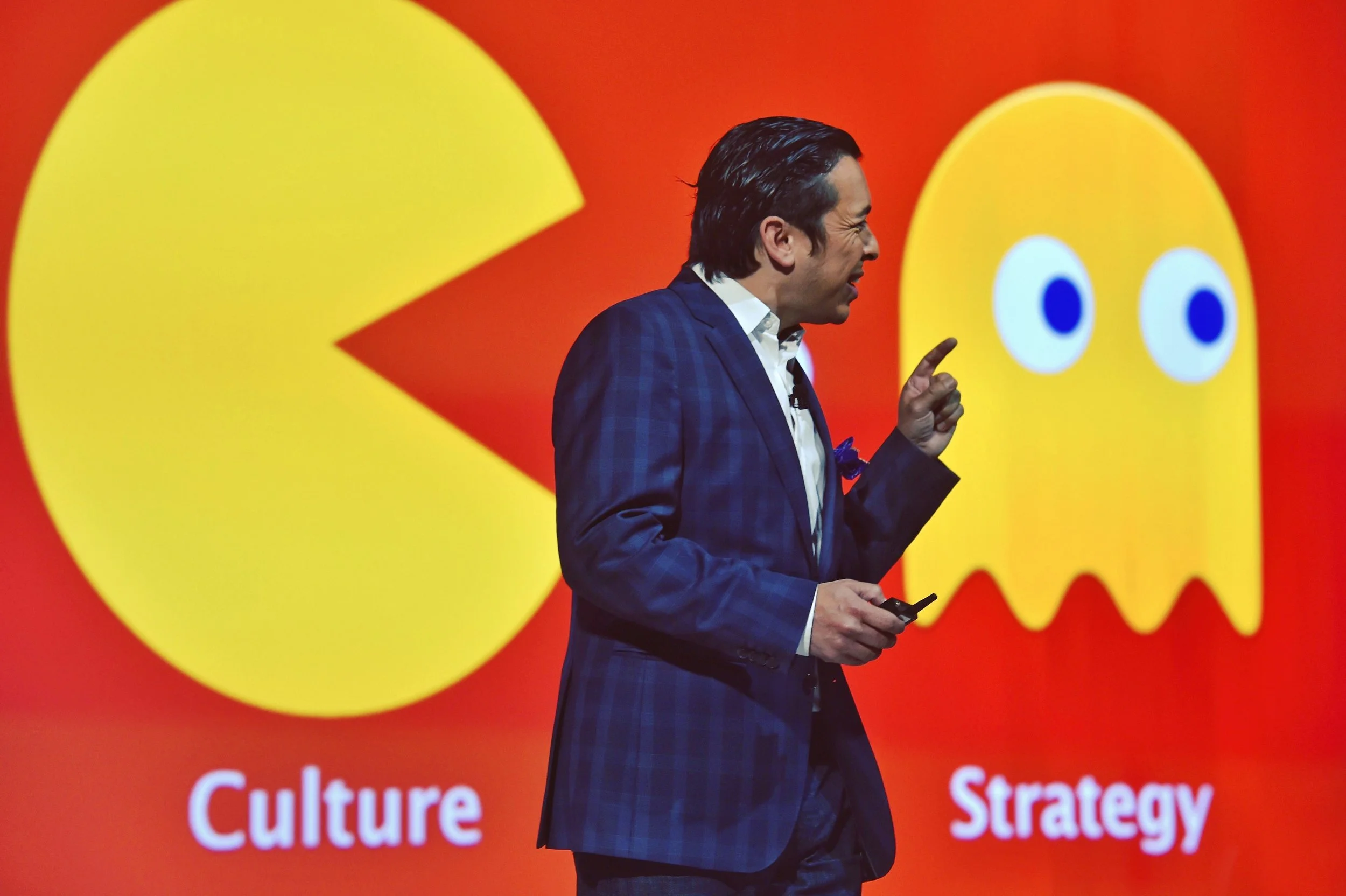 Un homme en costume parlant lors d'une présentation, avec un arrière-plan rouge comportant un symbole couleur Pac-Man et des mots en français et anglais comme 'Culture' et 'Strategy'.
