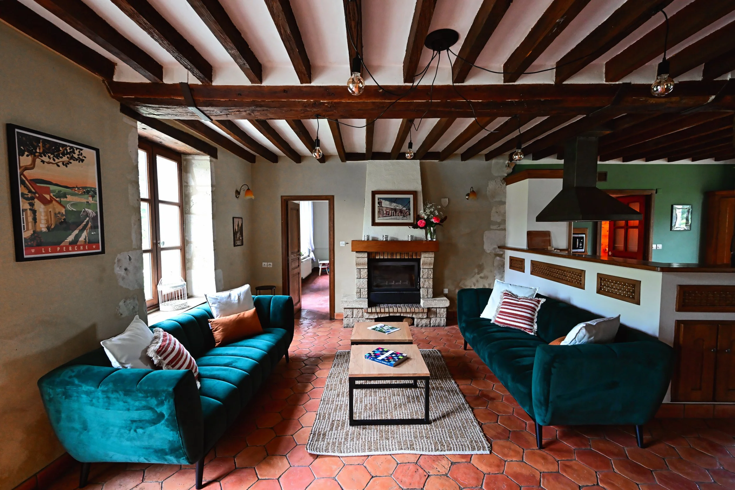 Intérieur d'un salon avec un plafond en poutres en bois, un manteau de cheminée en pierre, deux canapés verts avec des coussins rayés, une table basse en bois, des tableaux sur le mur, des fenêtres laissant entrer la lumière naturelle, et un sol en c