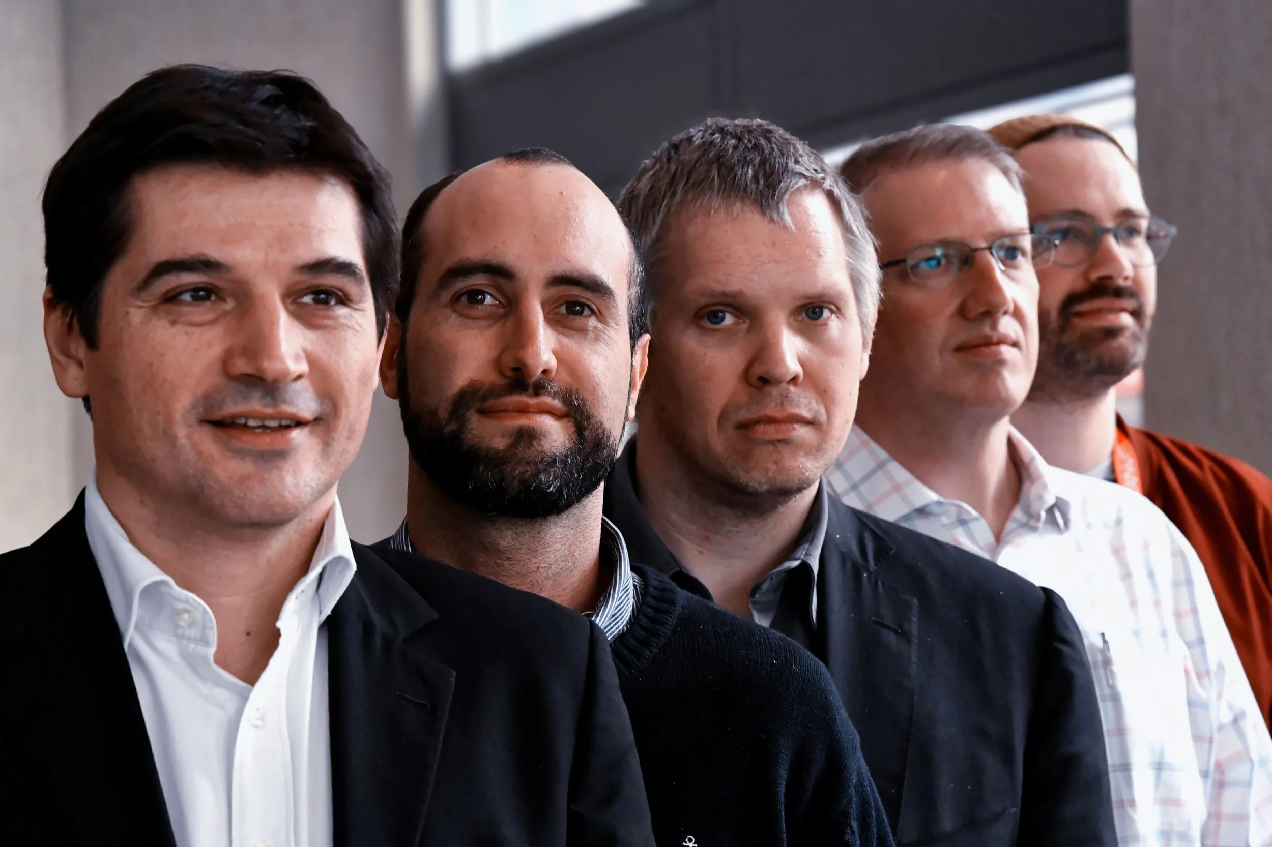 Groupe de cinq hommes debout côte à côte regardant vers l'avant, en intérieur, portant des vêtements professionnels, certains avec des lunettes.