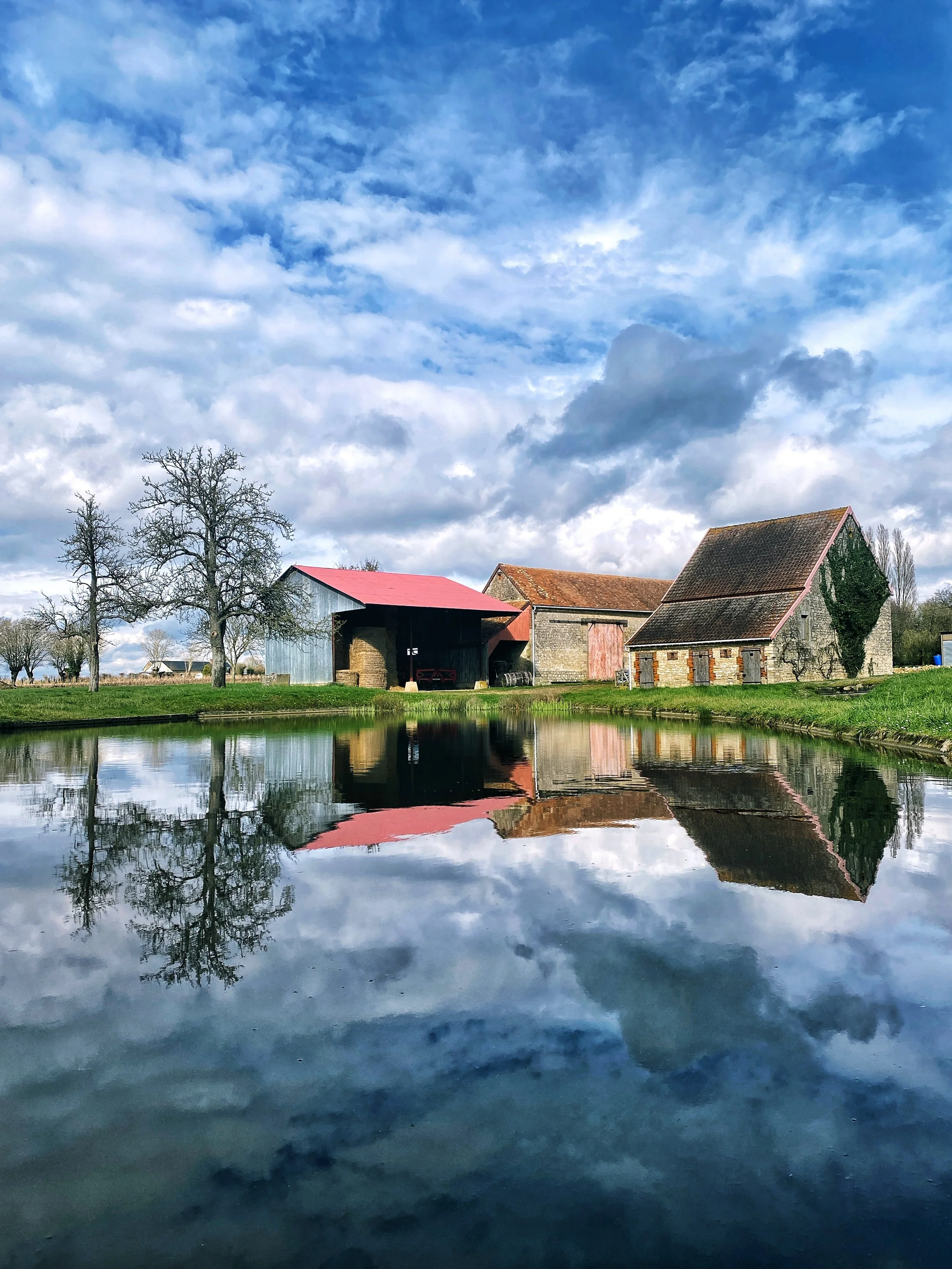 Paysage rural avec des maisons en pierre, un arbre nu, un étang reflétant le ciel orageux avec des nuages gris et bleus.