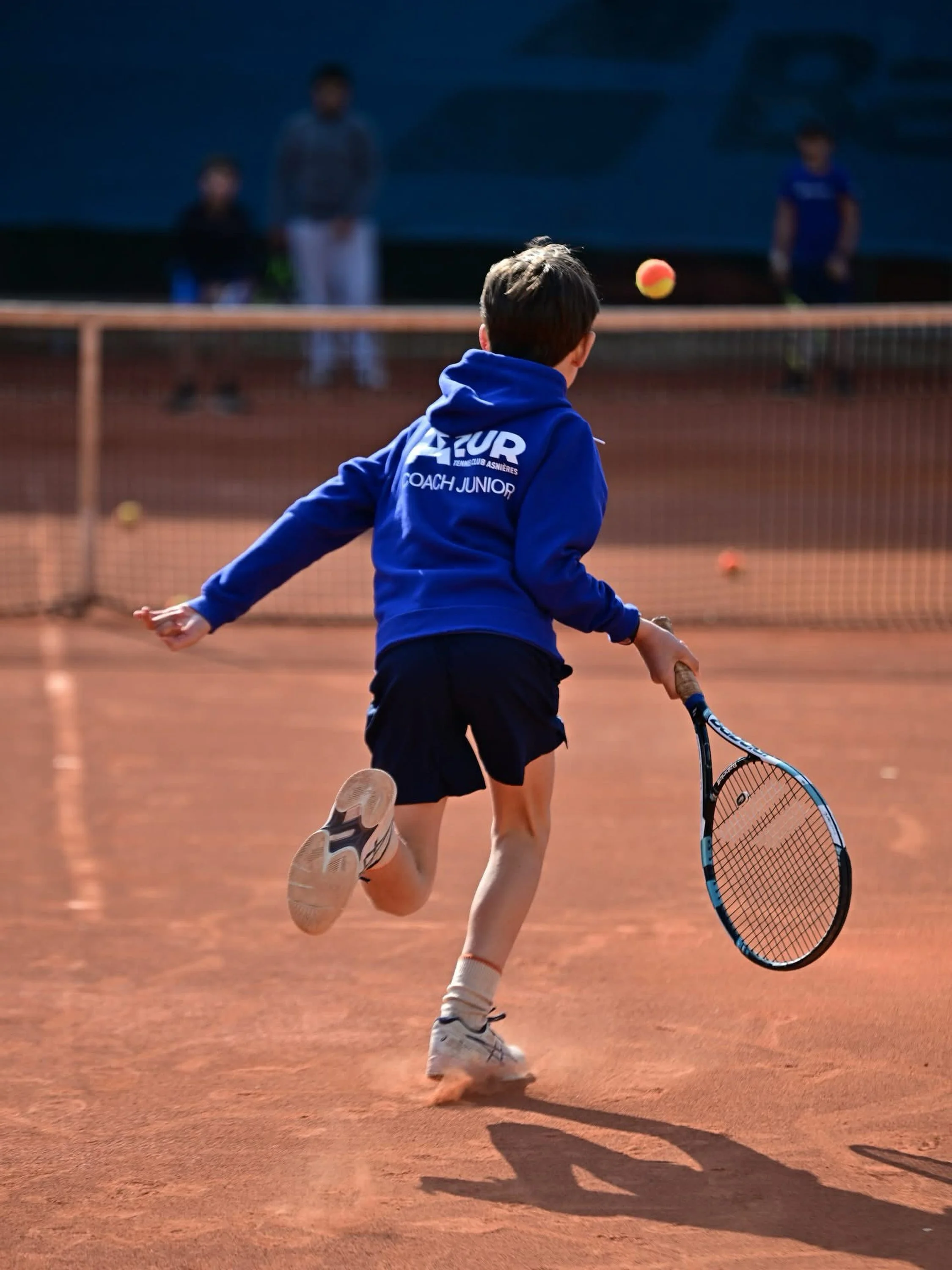 Un garçon joué au tennis sur un court en terre battue, portant un sweat à capuche bleu avec l'inscription 'AZUR Tennis Club Asnières' et 'Coach Junior'. Il court vers la balle de tennis en farandole, avec ses amis à l'arrière-plan.