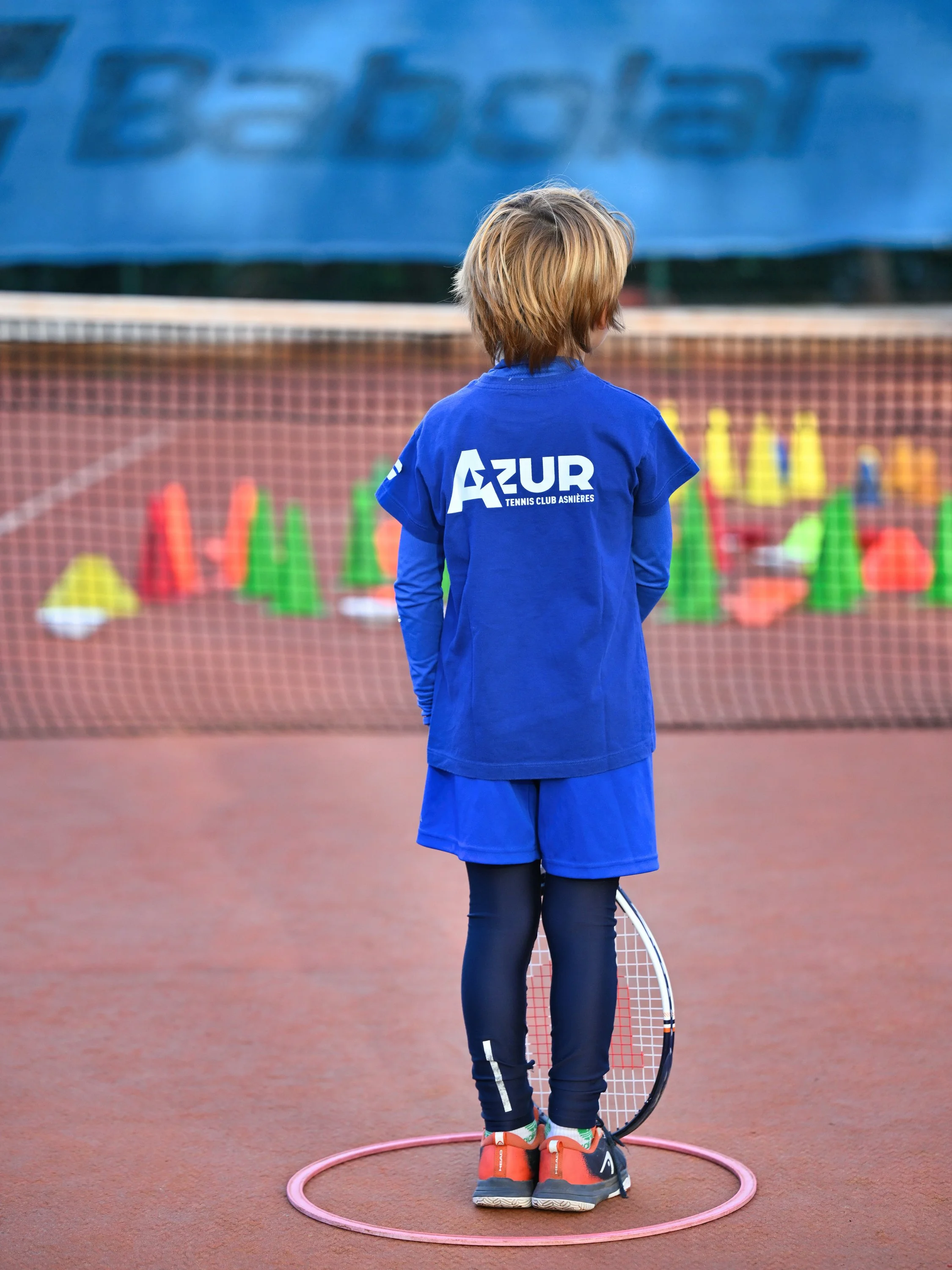 Un jeune garçon sur un court de tennis, vu de dos, portant un maillot bleu avec le logo 'AZUR Tennis Club Asnières' et tenant une raquette, entouré d'un cercle rose sur la surface.