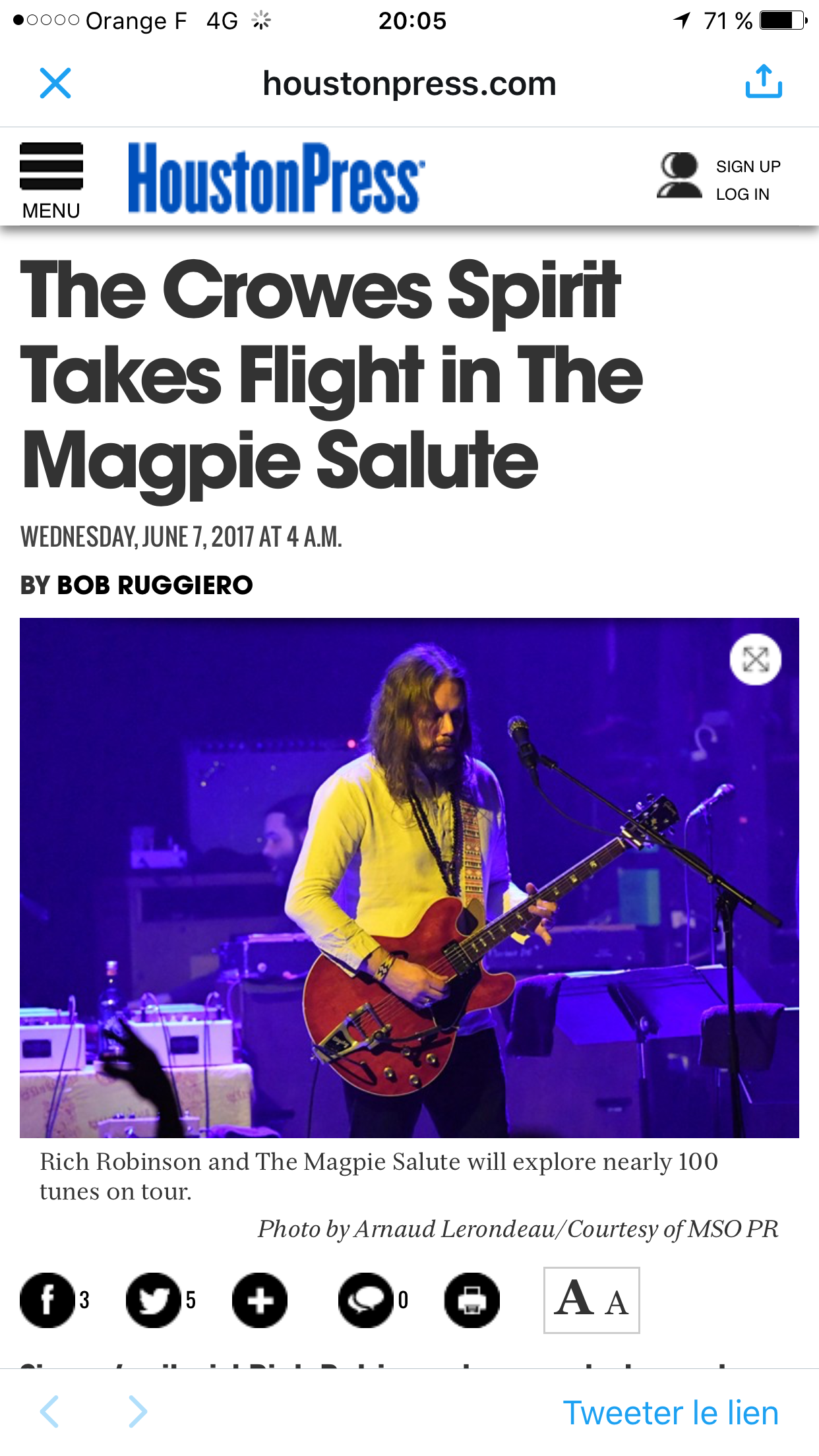 Un homme joue de la guitare électrique sur scène, éclairé en violet, avec d'autres instruments en arrière-plan et un microphone. Il a les cheveux longs et porte une chemise claire.