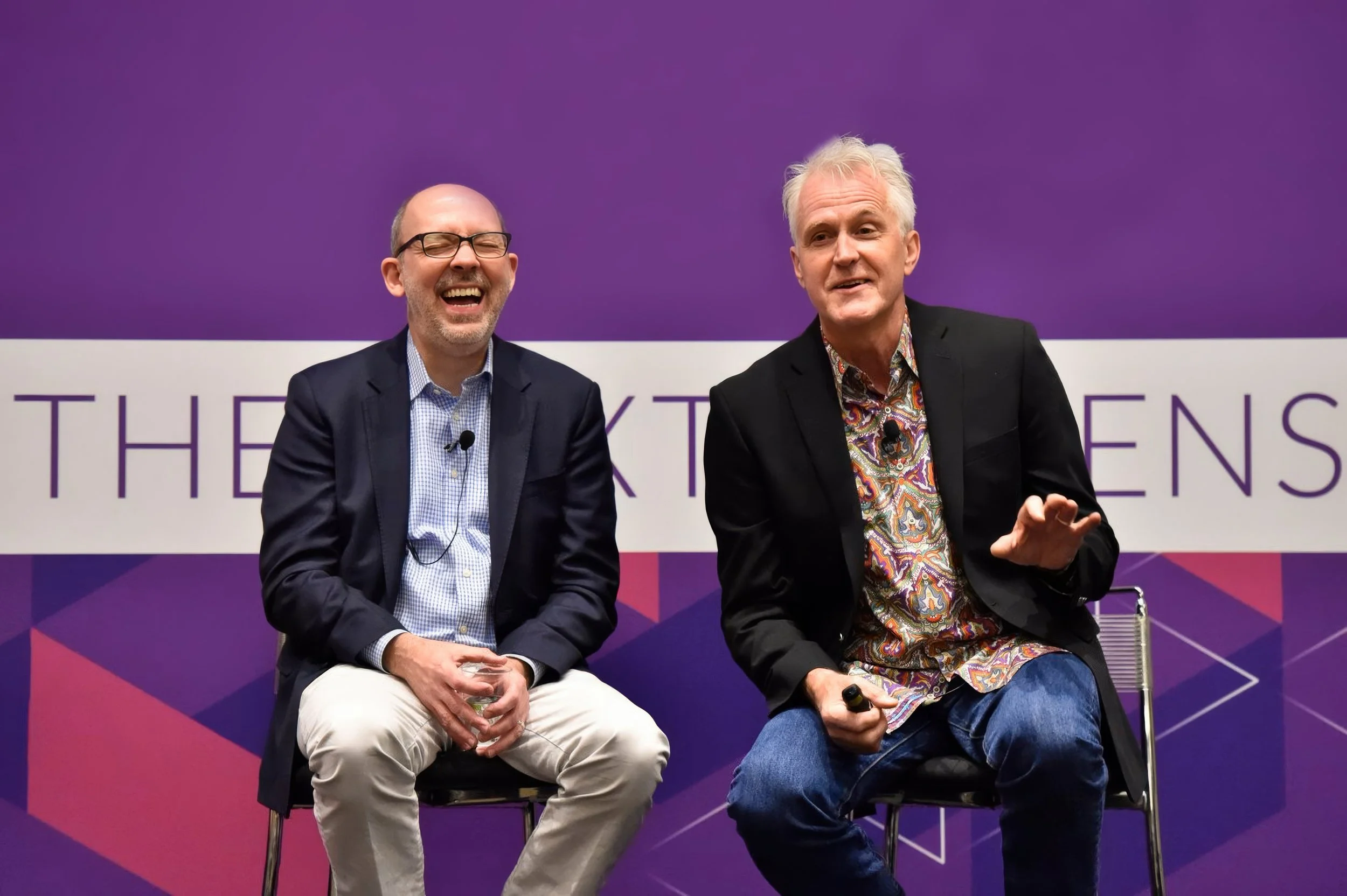 Deux hommes assis sur scène, en discussion, avec un fond violet et des lignes géométriques. L'un porte un costume sombre avec une chemise à carreaux, l'autre un blazer noir avec une chemise à motifs colorés.