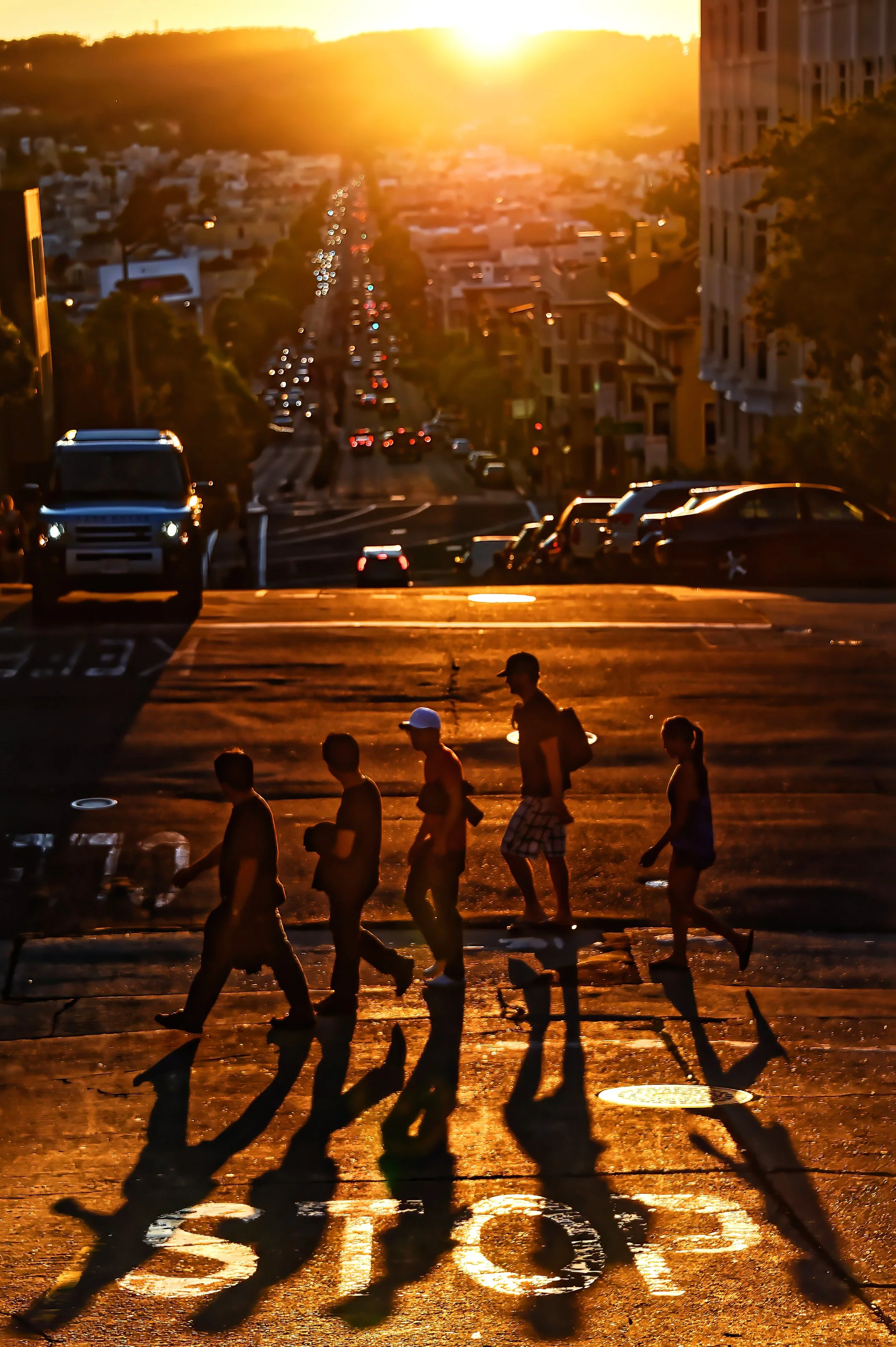 Cliché d'une rue en fin d'après-midi ou début de soirée avec des personnes qui traversent un passage piéton sous un soleil couchant, avec des voitures en circulation et un bâtiment résidentiel à droite.