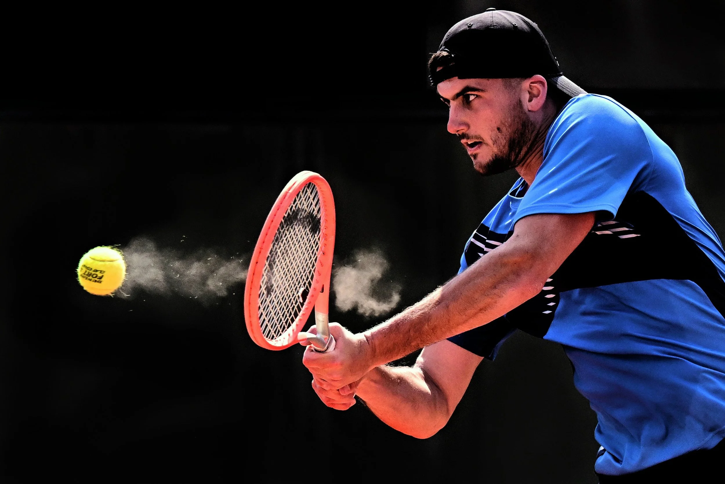 Un homme jouant au tennis, en train de frapper la balle avec une raquette, portant un maillot bleu et une casquette noire, fond sombre.