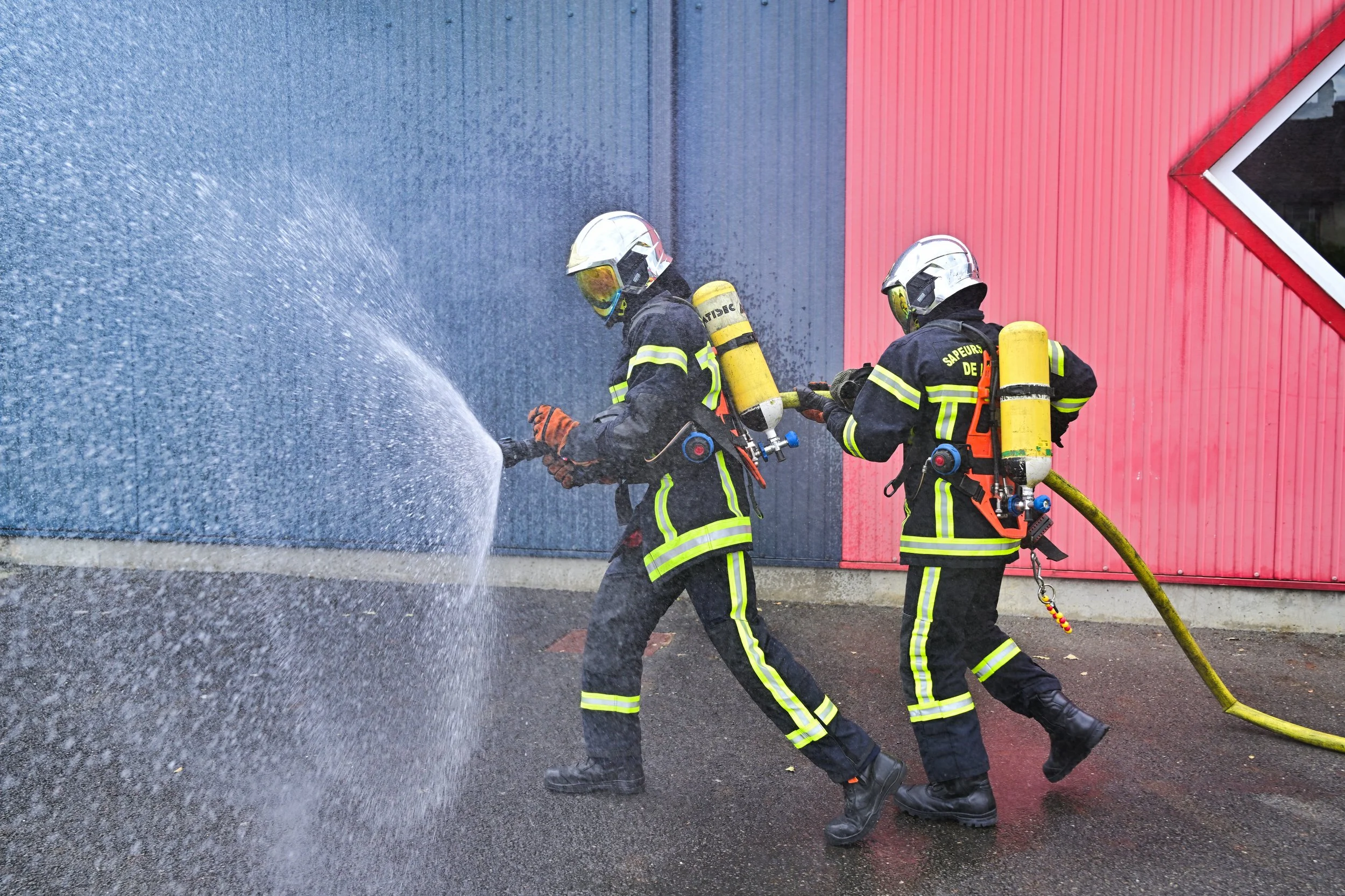 Deux pompiers en équipement de protection, utilisant une lance à eau pour éteindre un incendie sur un bâtiment avec une façade rouge et bleue.