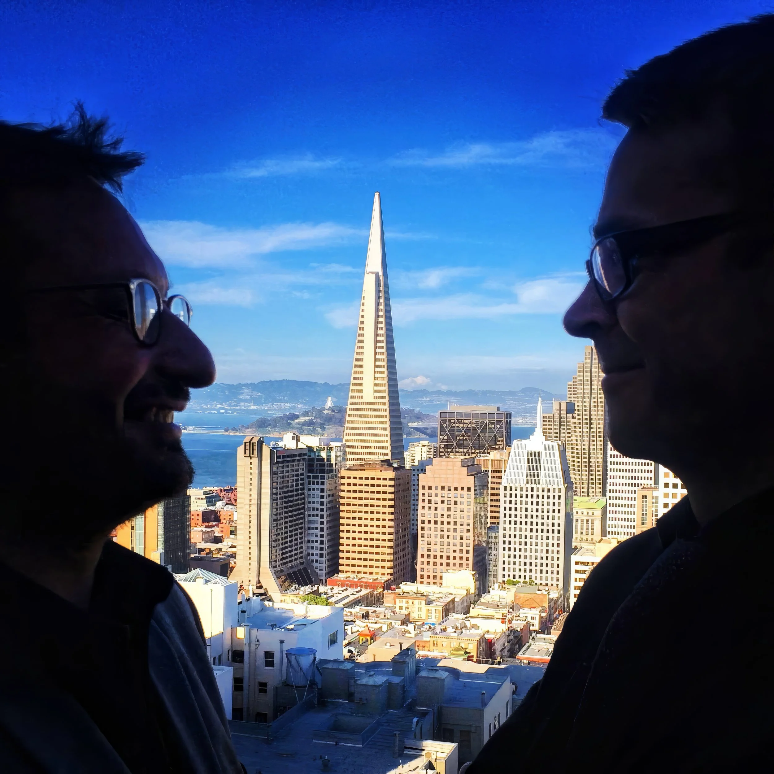 Deux personnes en profil regardent vers un ciel bleu avec la ville de San Francisco en arrière-plan, notamment le Sutro Tower, une tour de télécommunications en forme de pyramide effilée.