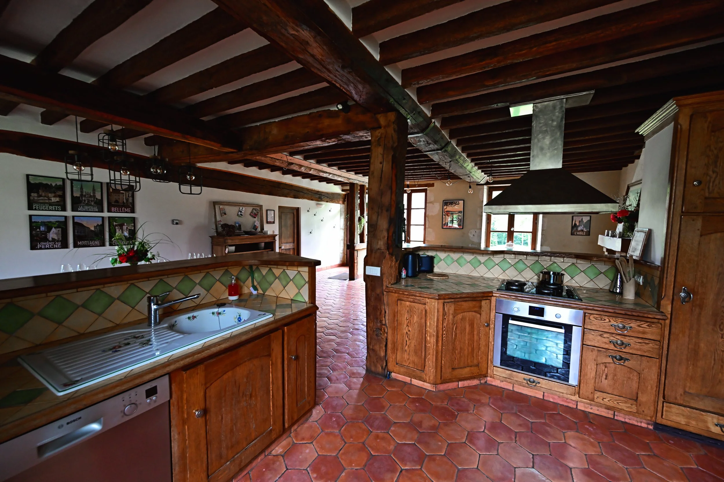Cuisine avec poutres en bois apparentes, plan de travail en carreaux verts et orangés, évier en porcelaine, four encastré, notamment une scène intérieure chaleureuse et rustique.