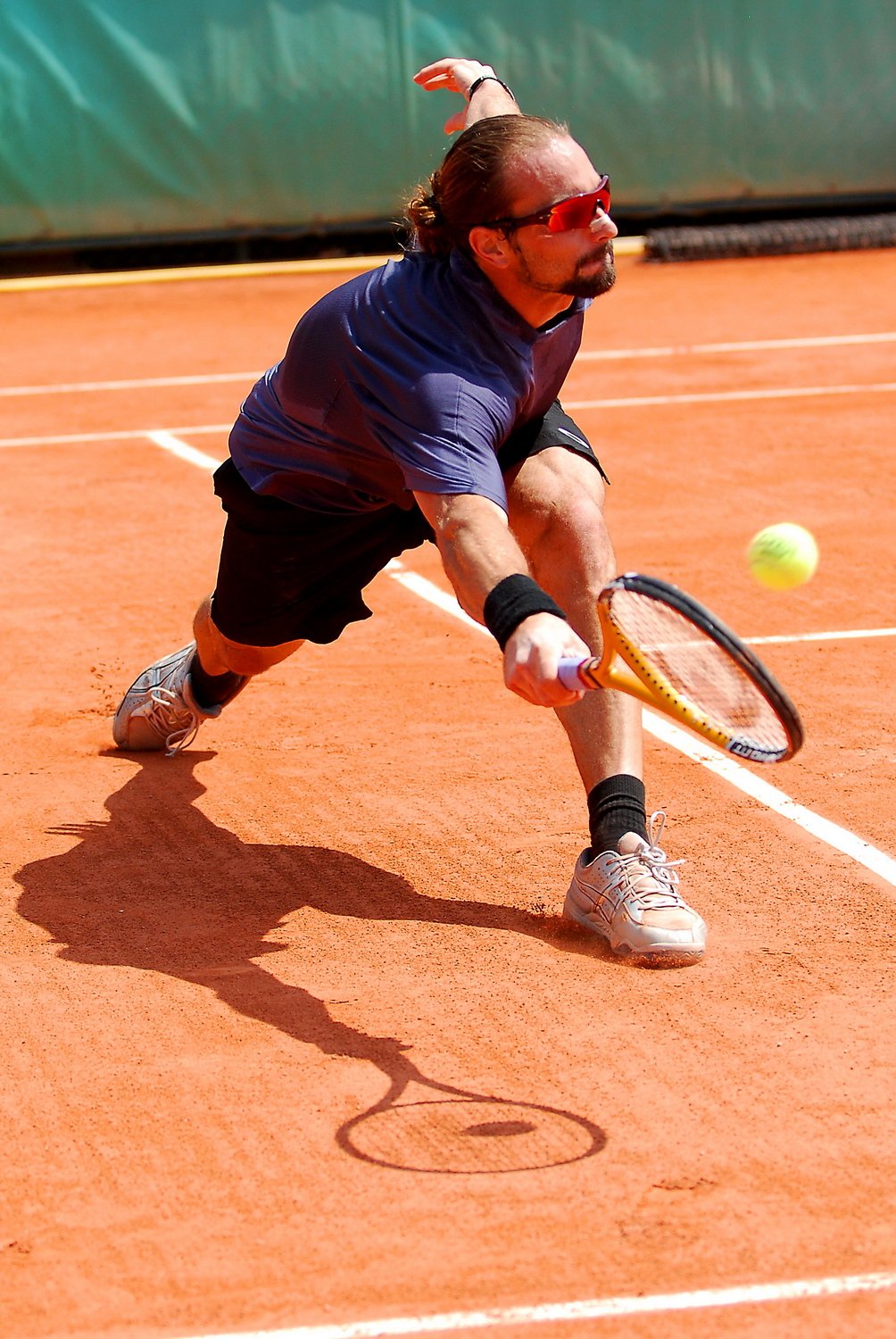 Un homme joue au tennis sur un court en terre battue, s'apprêtant à frapper une balle de tennis avec une raquette. Il porte des lunettes de soleil, un t-shirt bleu, des chaussures de sport, des manchons autour des bras et est en position accroupie