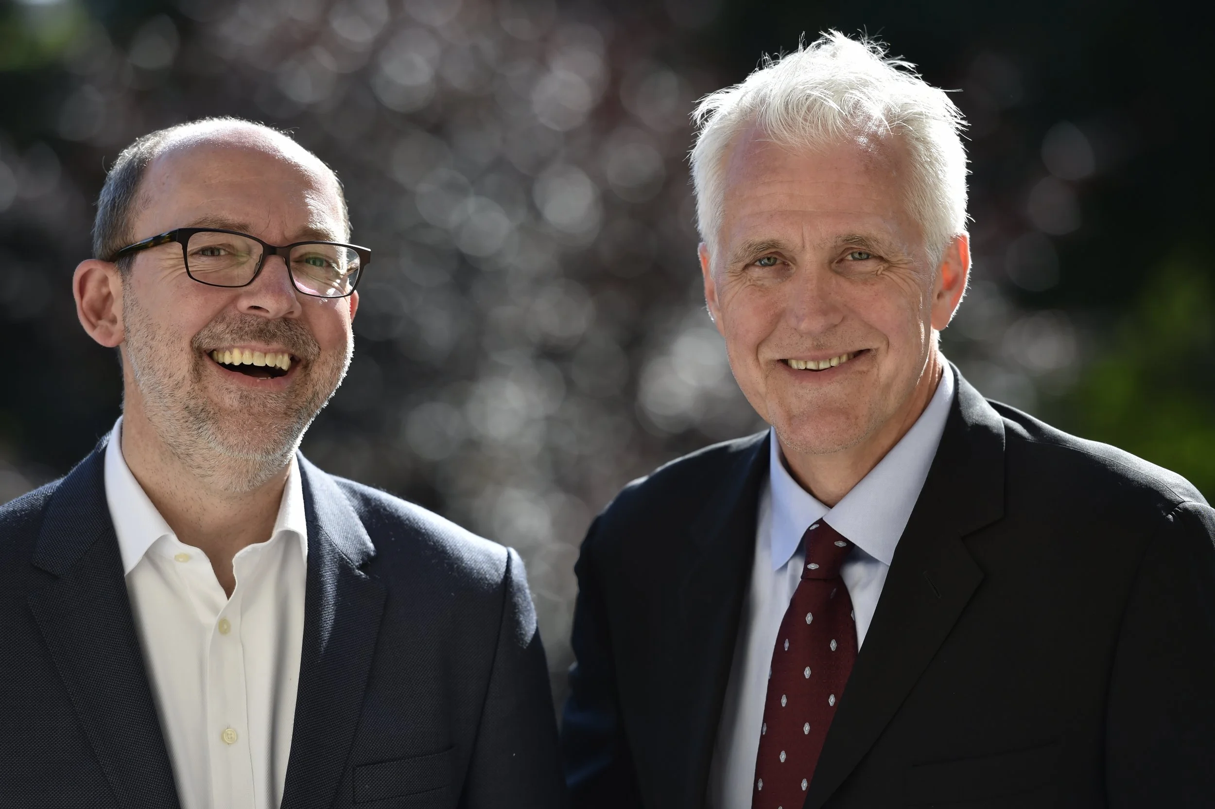 Deux hommes d'affaires souriants portant des costumes, l'un avec des lunettes, en plein air avec un arrière-plan flou de nature.