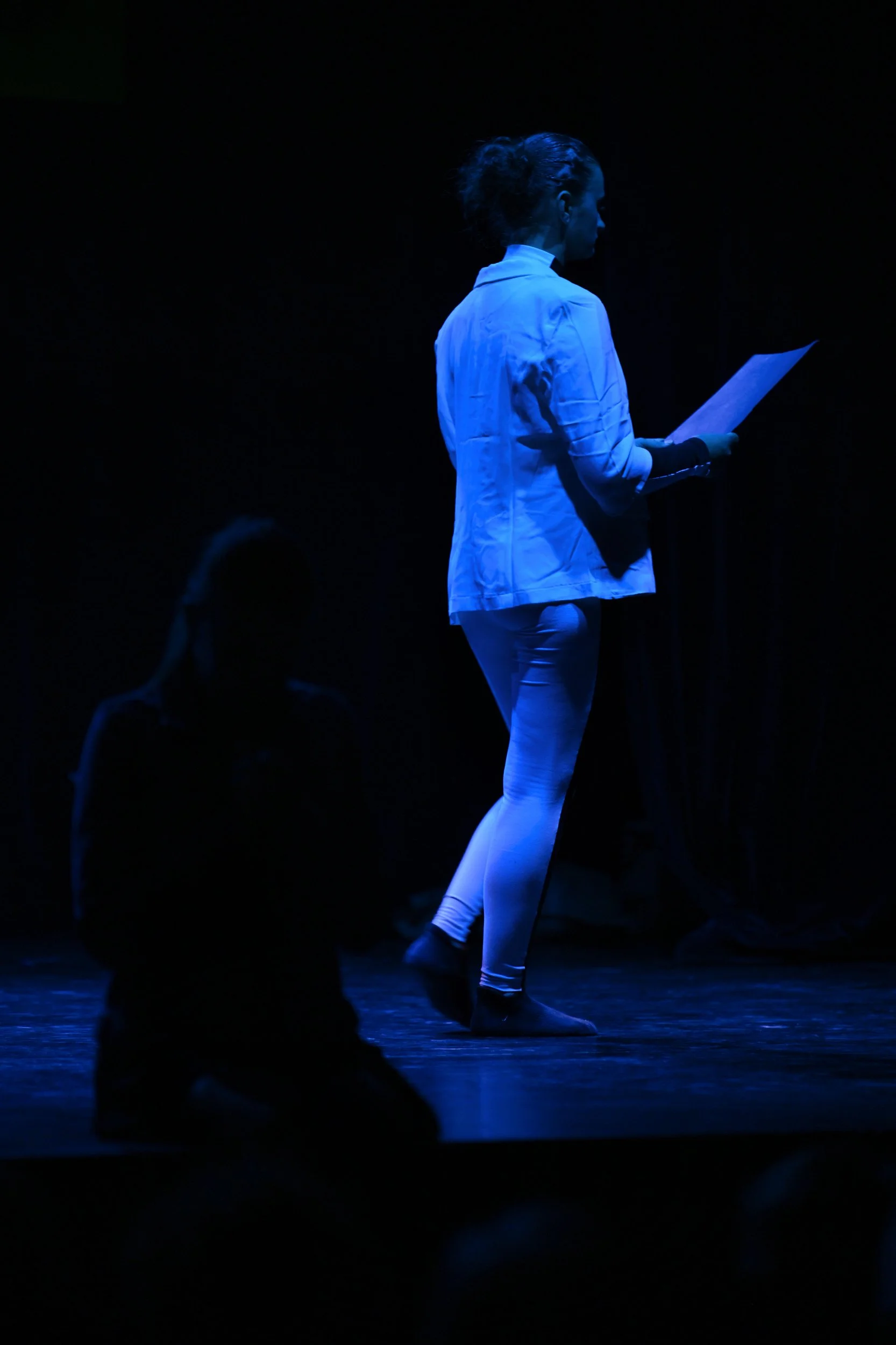 Une femme en blanc, vue de profil, lit un texte sur une feuille dans une scène sombre avec un éclairage bleu. Deux autres personnes sont assises dans l'ombre dans le fond.