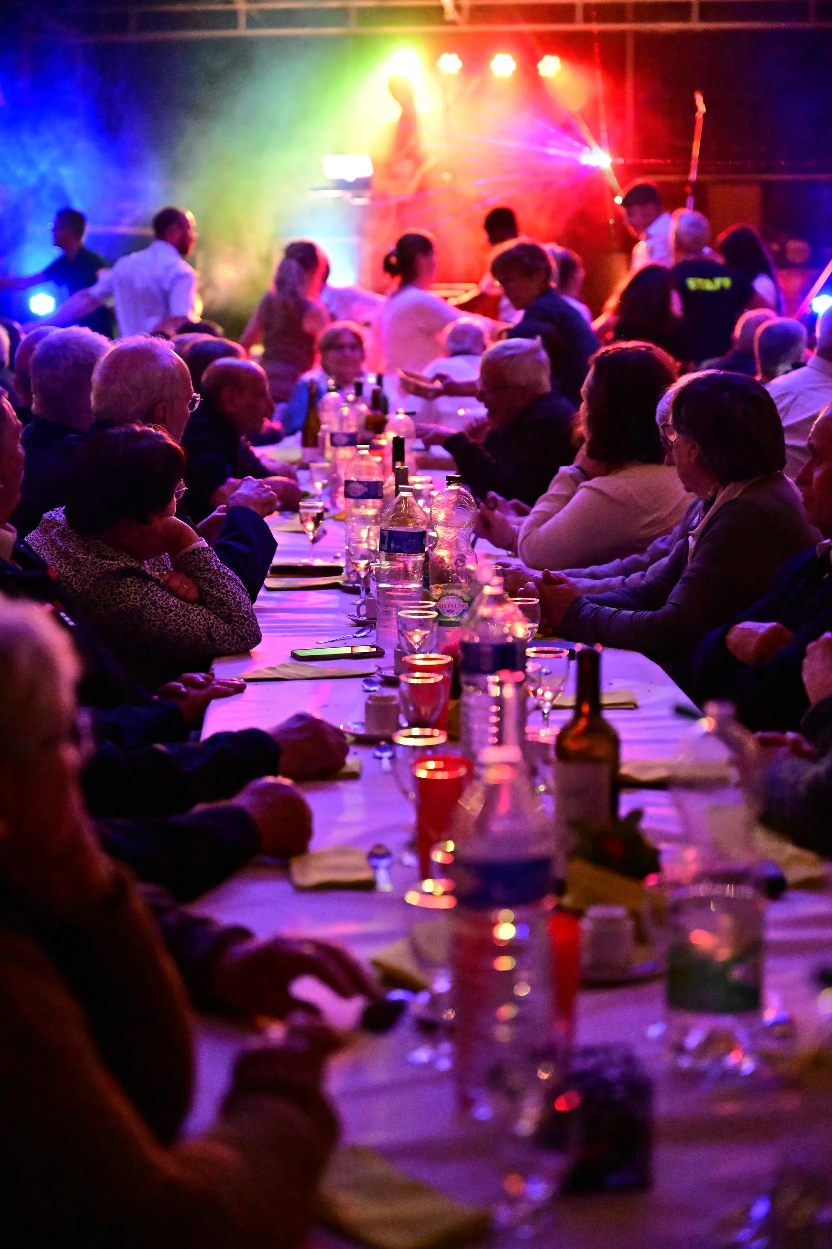 Une grande salle où de nombreuses personnes sont assises à une longue table lors d'un repas ou d'une célébration. La scène est illuminée de lumières colorées, avec des couleurs comme le rouge, le jaune, le vert, et le violet, créant une ambiance fest
