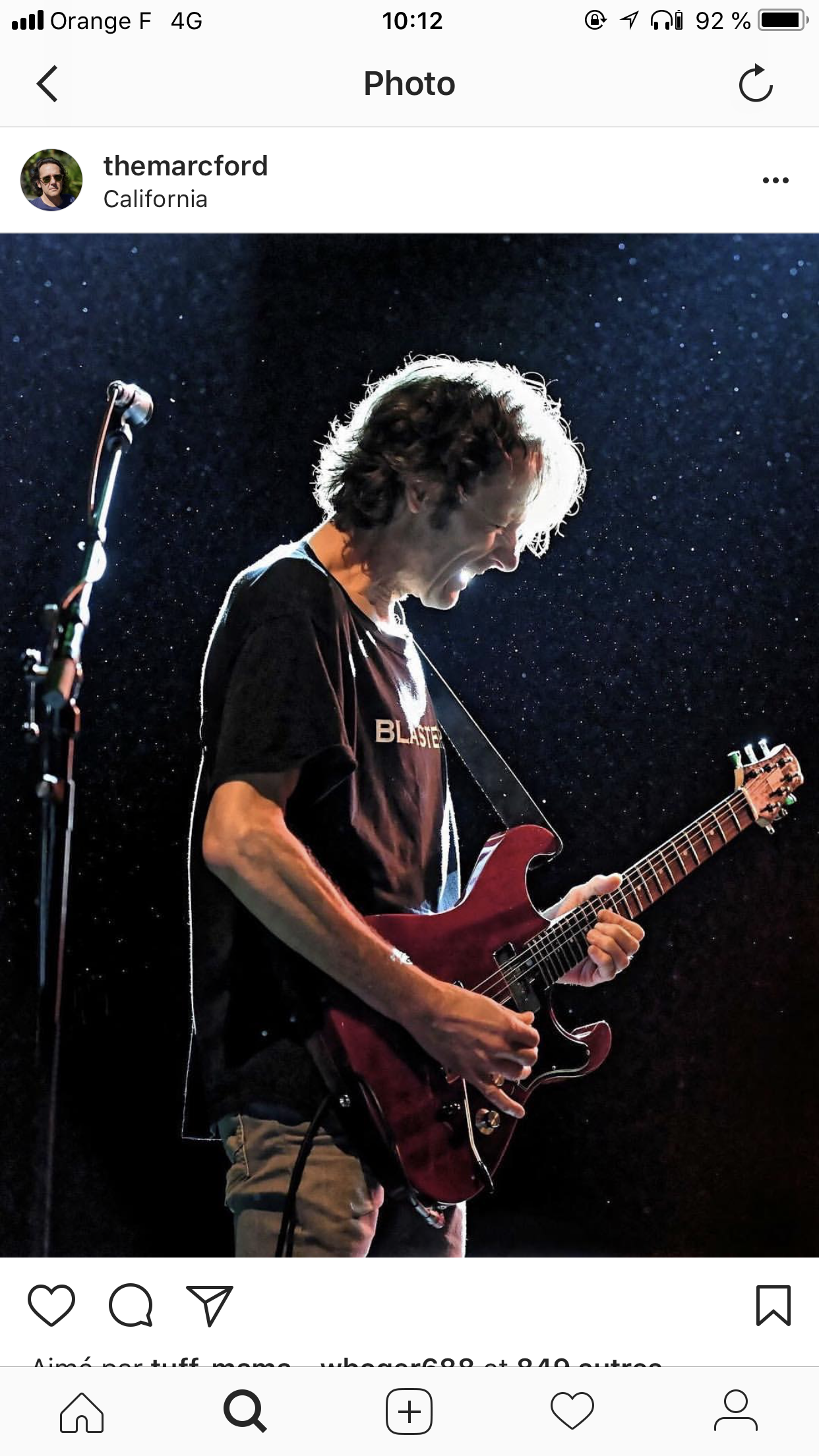 Un homme joue de la guitare électrique sur scène, éclairé par un éclairage brillant contre un fond sombre avec des points lumineux, en train de sourir ou de chanter.