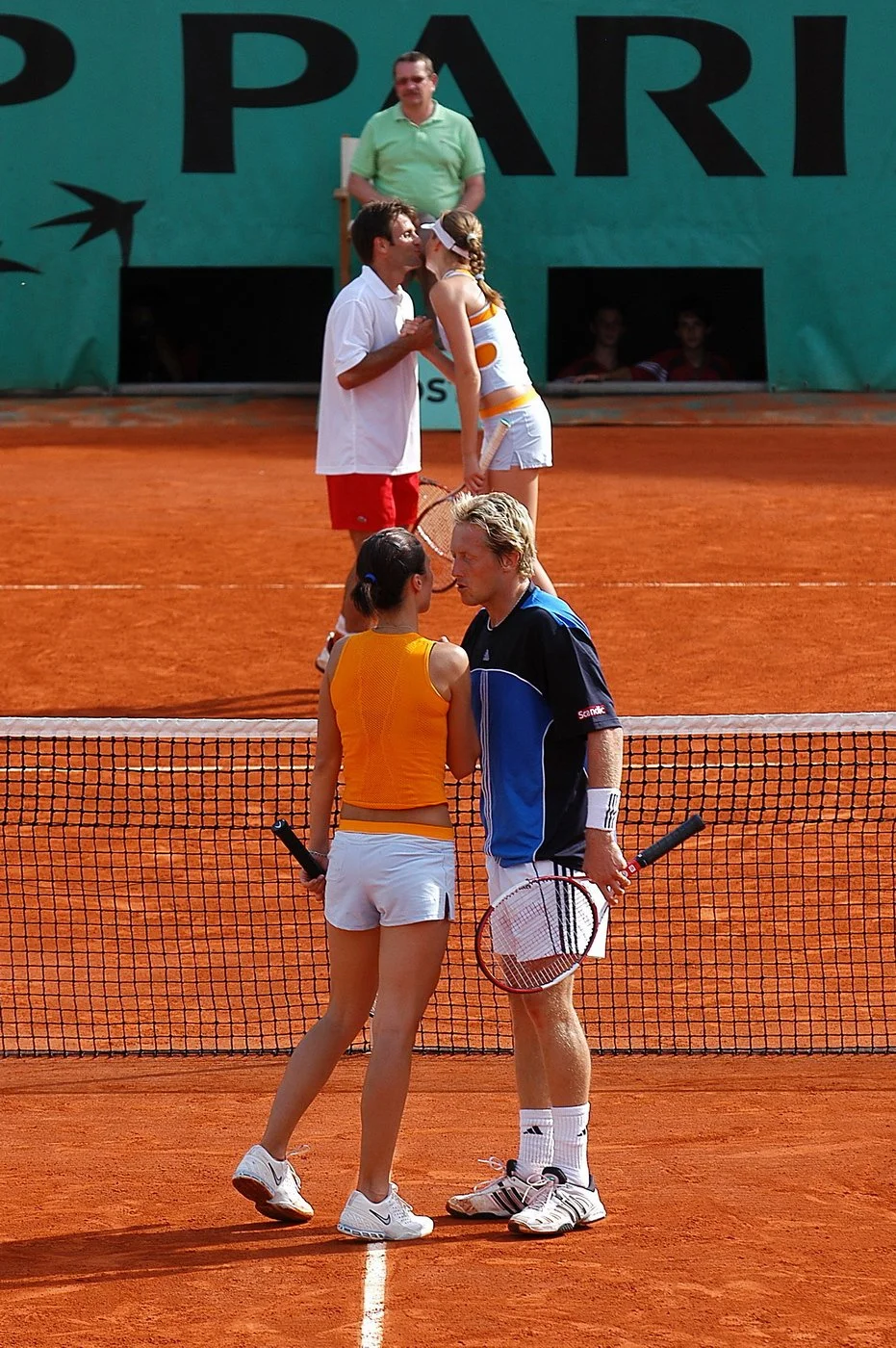 Deux joueurs de tennis, une femme en tenue orange et une homme en tenue noire et blanche, discutent près du filet sur un court en terre battue. Deux autres joueurs, un homme en vert et une femme en blanc et orange, se tiennent derrière eux, avec un h
