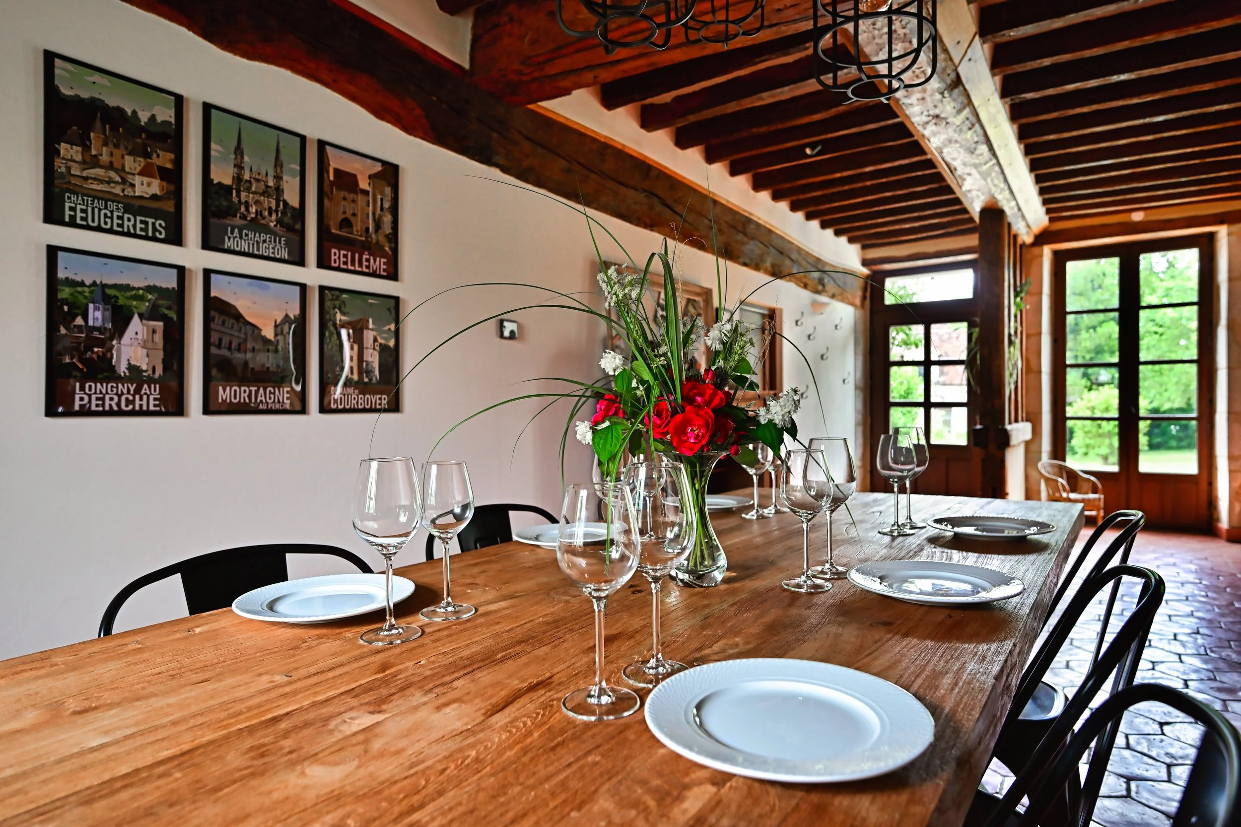 Intérieur d'une salle à manger avec un long plan de table en bois, décorée d'une vase avec des fleurs rouges et blanches, entourée de verres à vin et d'assiettes blanches, avec des murs décorés de posters de lieux touristiques et une grande fenêtre l