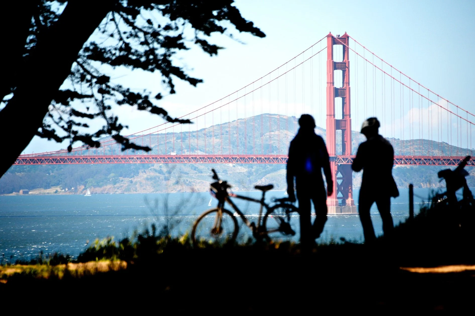 Silhouettes de deux personnes et un vélo près de l'eau, avec le pont Golden Gate en arrière-plan dans le sud de la baie de San Francisco, sous un ciel clair.