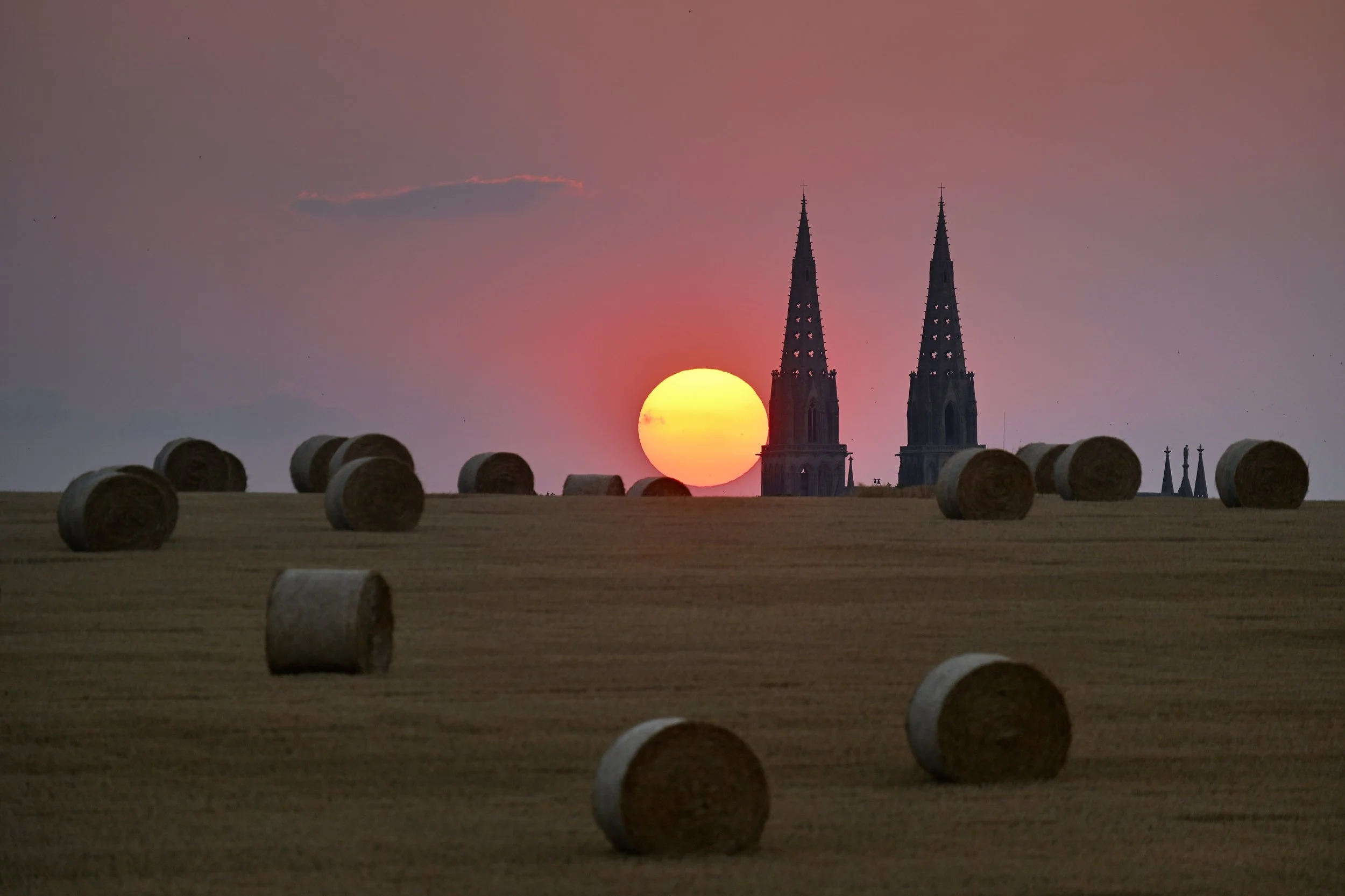 Coucher de soleil derrière deux clochers d'église avec des ballots de foin en premier plan.