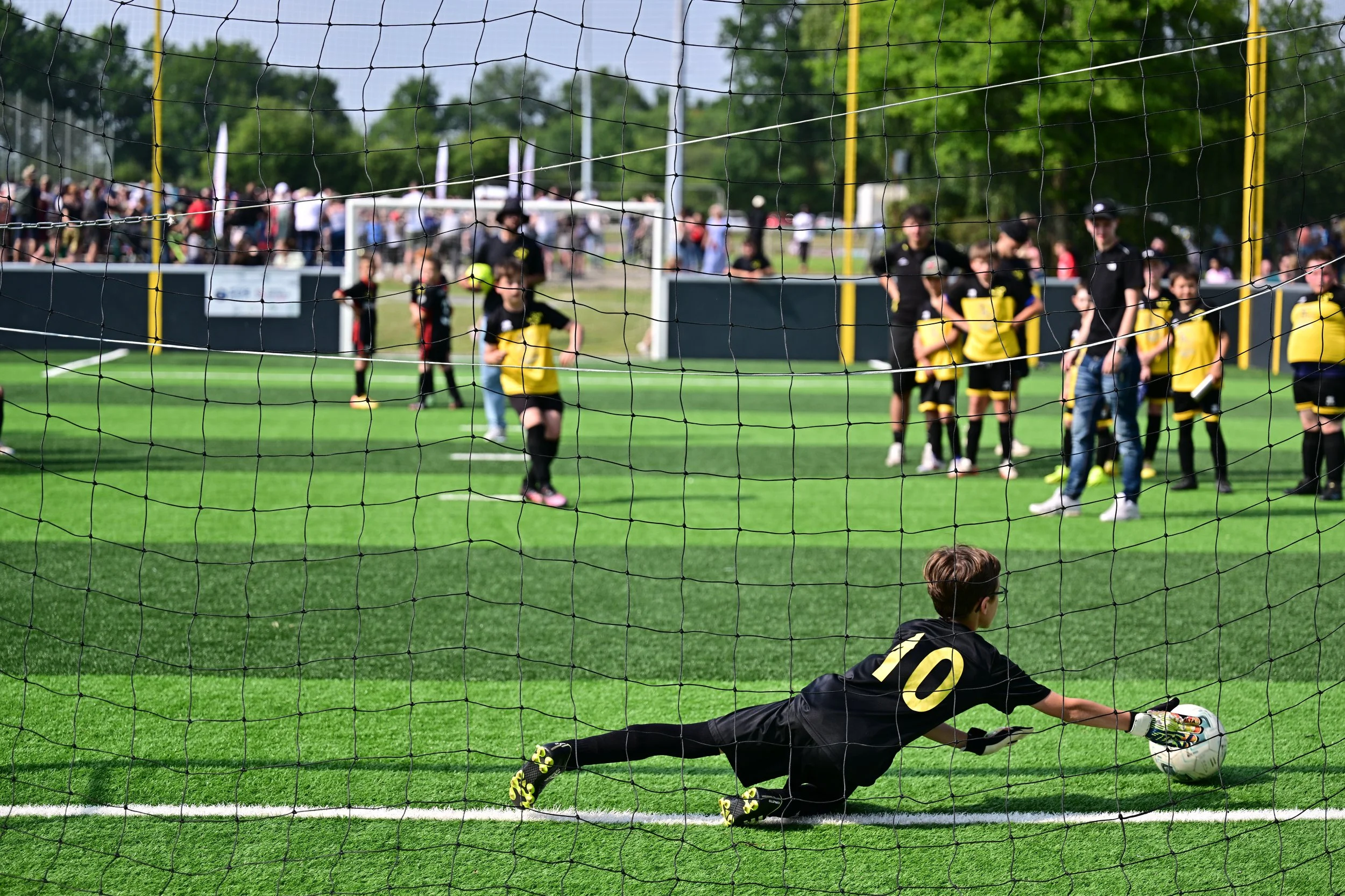 Un gardien de but en maillot noir et jaune effectue un arrêt lors d'un match de football, avec un groupe de joueurs et un public en arrière-plan, vu à travers un filet de but.