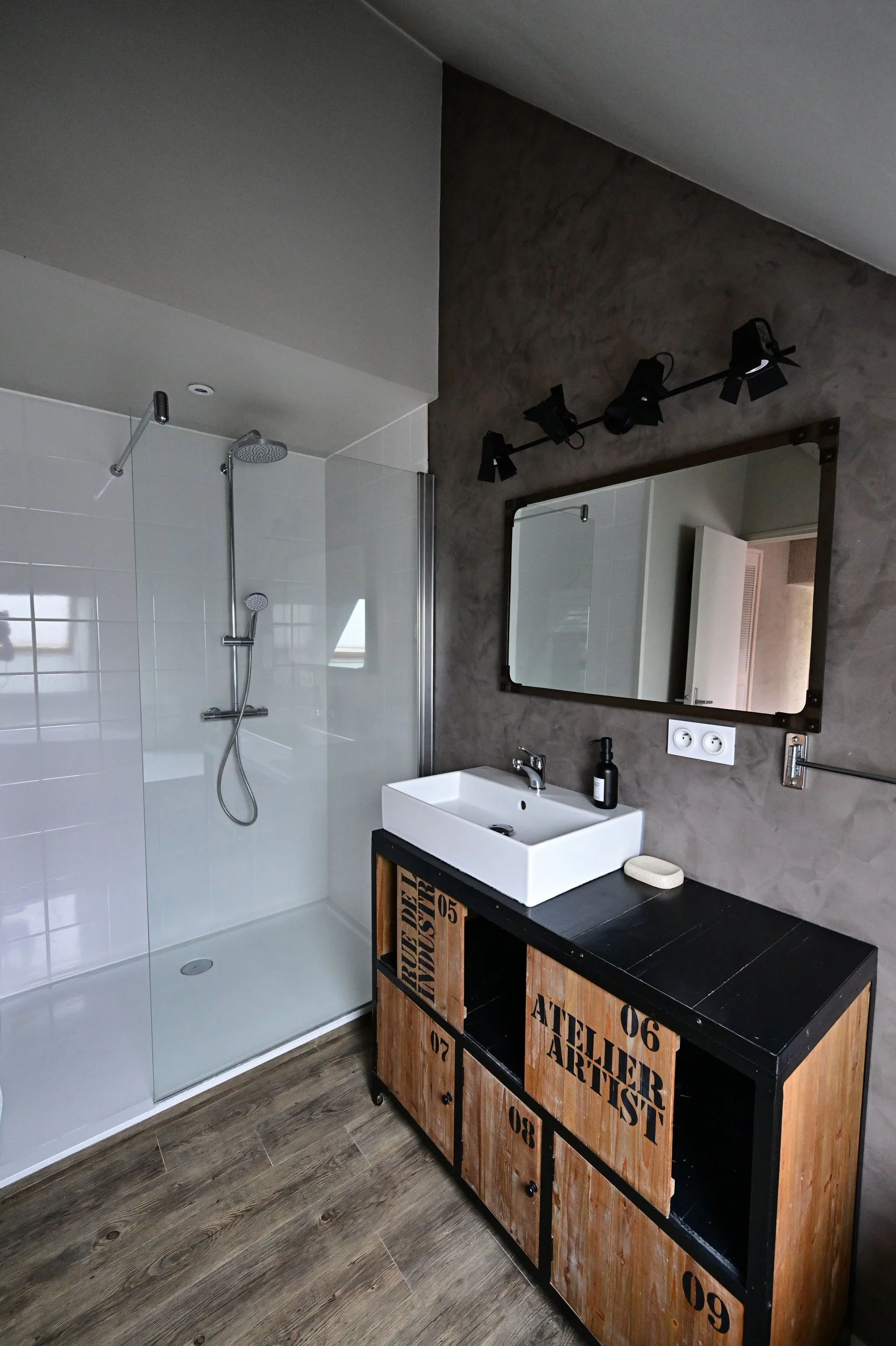 Salle de bain moderne avec une douche à l'italienne, un miroir rectangulaire au mur, un lavabo blanc sur une console en bois noir, un meuble en bois avec des tiroirs numérotés, et un éclairage noir au plafond.