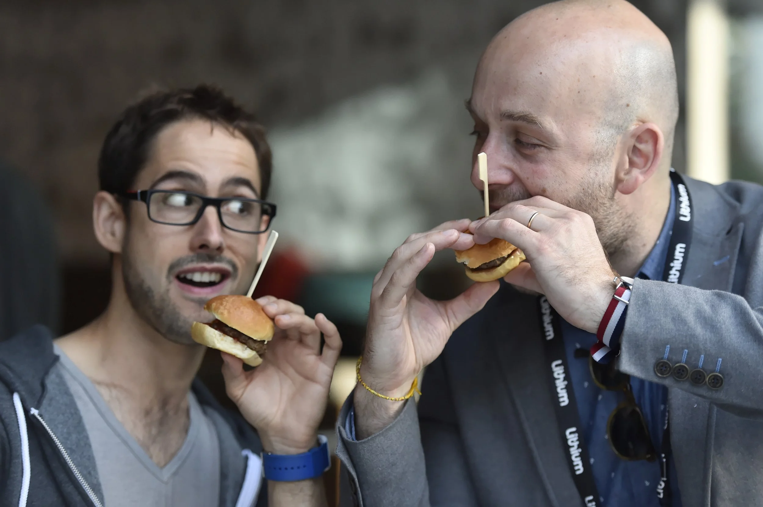 Deux hommes se regardent en mangeant des hamburgers, l'un porte des lunettes noires et un sweat gris, l'autre un blazer gris avec une écharpe et un bracelet aux couleurs françaises.