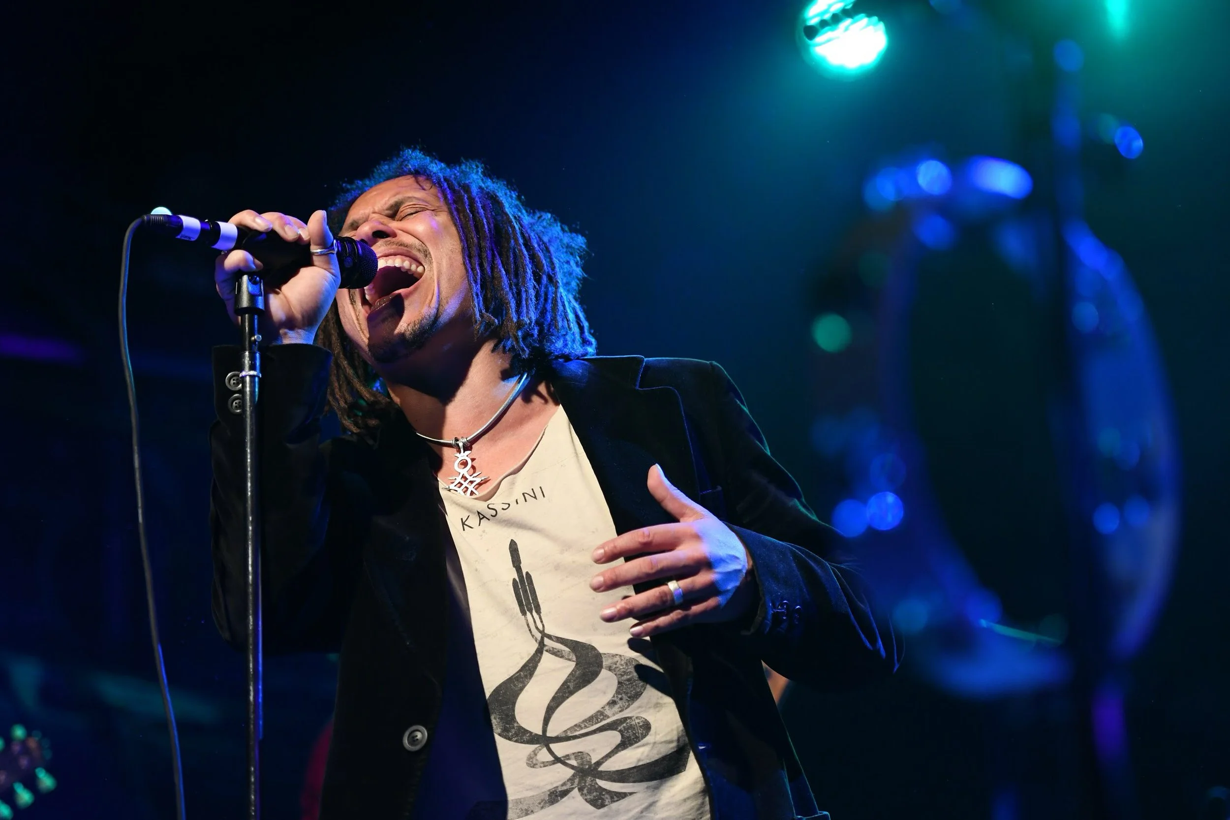 Un homme avec des dreadlocks noirs chante dans un microphone lors d'une performance scénique, portant un t-shirt blanc et une veste noire, avec un fond éclairé de lumières colorées.
