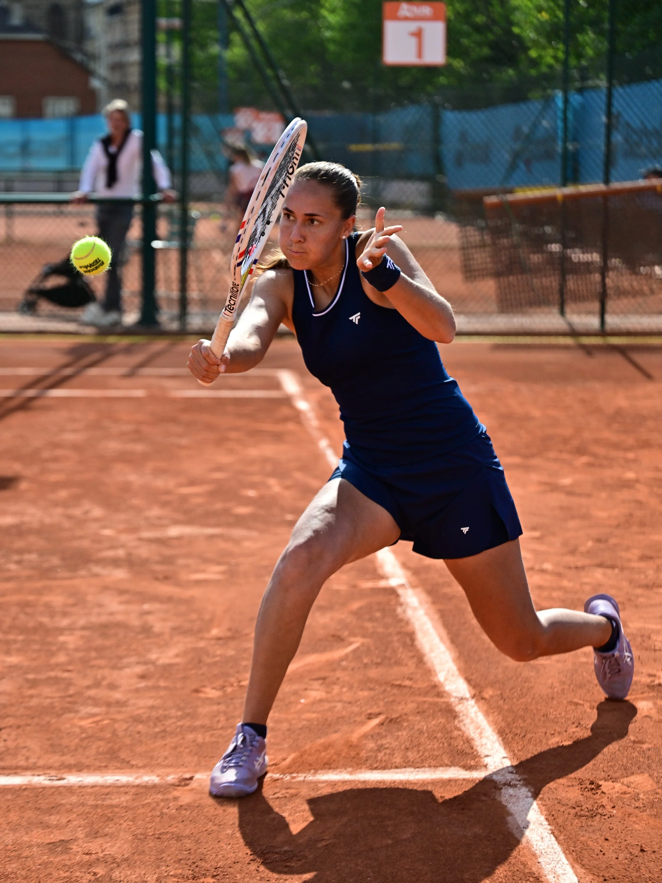 Une jeune femme joue au tennis sur un court en terre battue, en train de frapper une balle. Elle porte une tenue de sport bleue et des chaussures de tennis blanches. En arrière-plan, il y a un panneau indiquant le numéro 1.