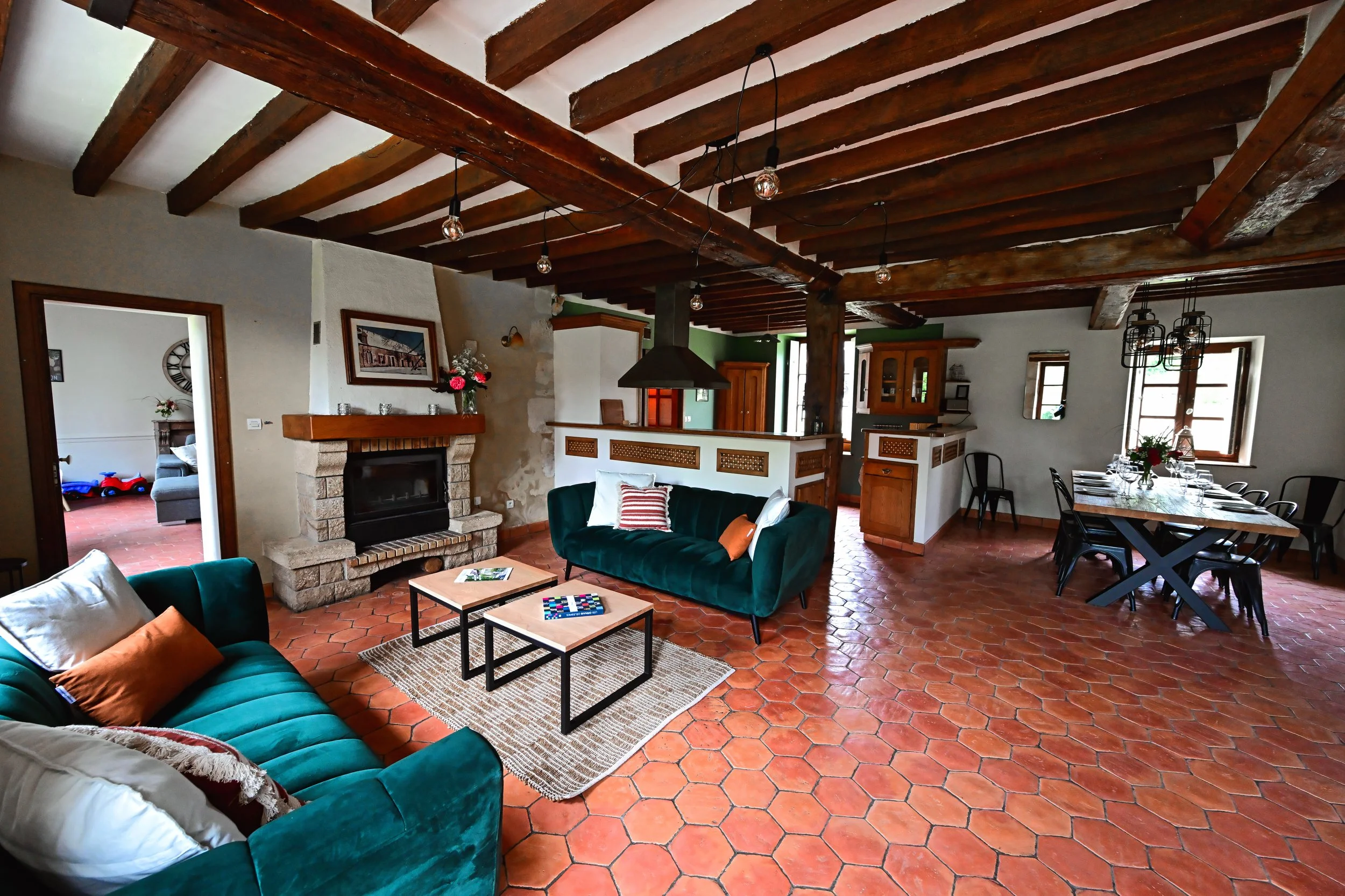 Salon avec poutres en bois au plafond, cheminée en pierre, canapé vert avec coussins, tables basses, mur blanc, fenêtres laissant entrer la lumière, et table à manger avec des chaises. Murs blancs et sol en carreaux de terre cuite.