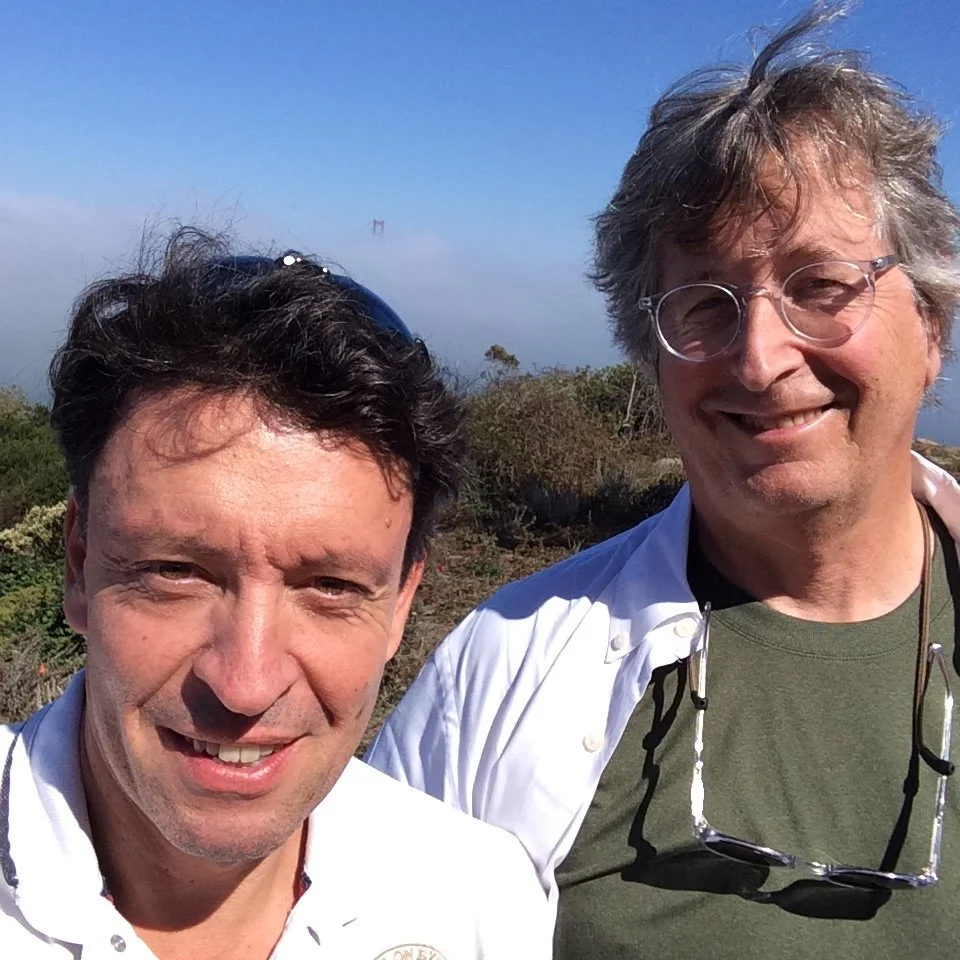 Deux hommes souriants prennent une photo à l'extérieur avec la Golden Gate Bridge en arrière-plan, enveloppée de brouillard, par une journée ensoleillée.