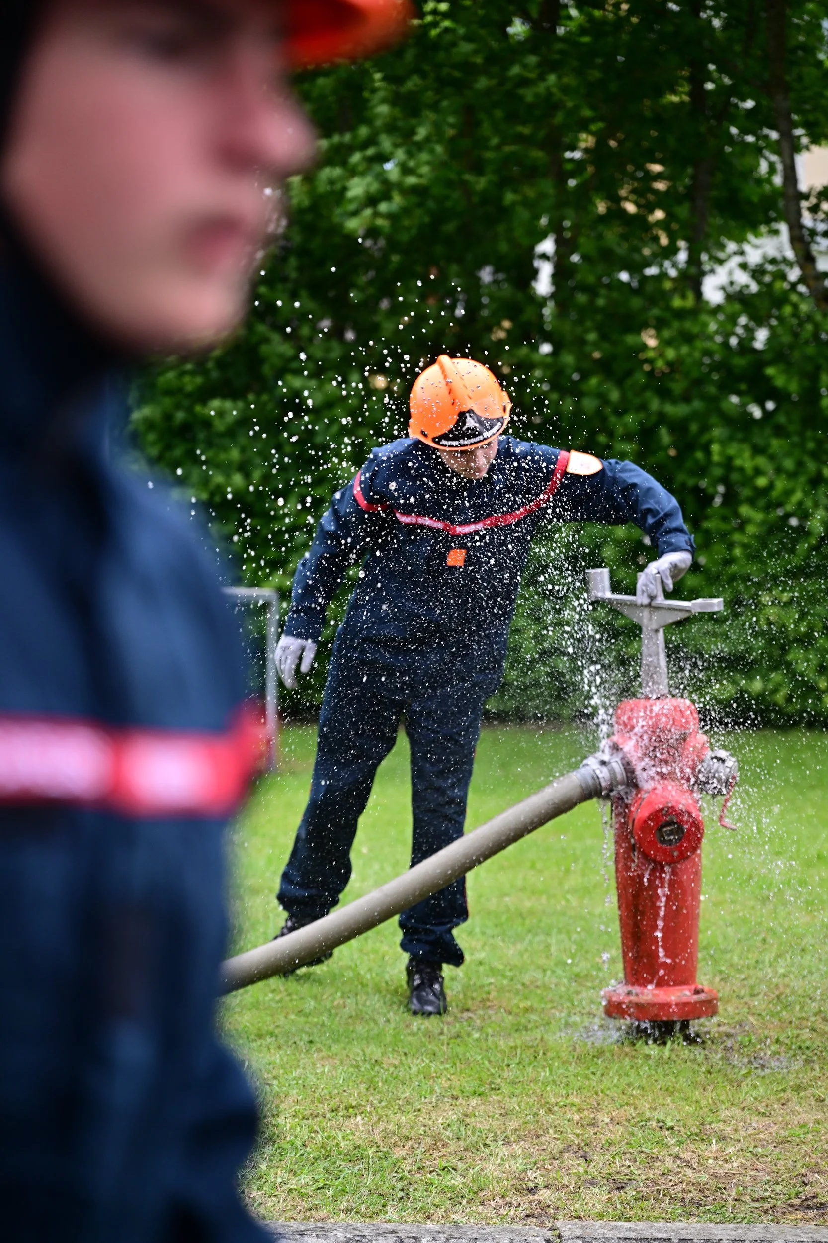 Deux pompiers en tenue de sécurité, portant un casque orange, interviennent avec un jet d'eau pour éteindre un incendie dans un espace vert, encadrés par des arbres.