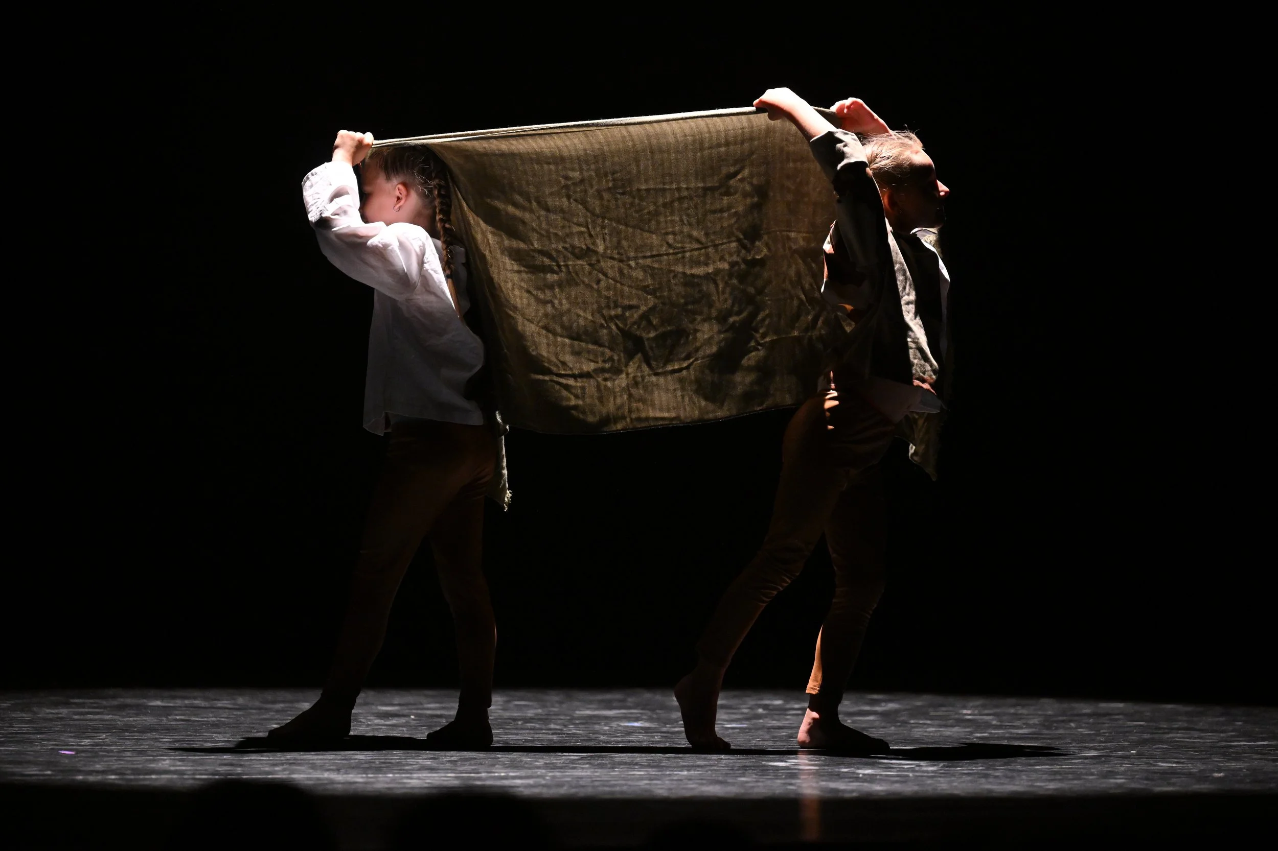 Deux danseurs en costume noir et blanc sur scène, portant une cape ou tissu beige, dans une lumière tamisée, créant un contraste avec l'arrière-plan sombre.