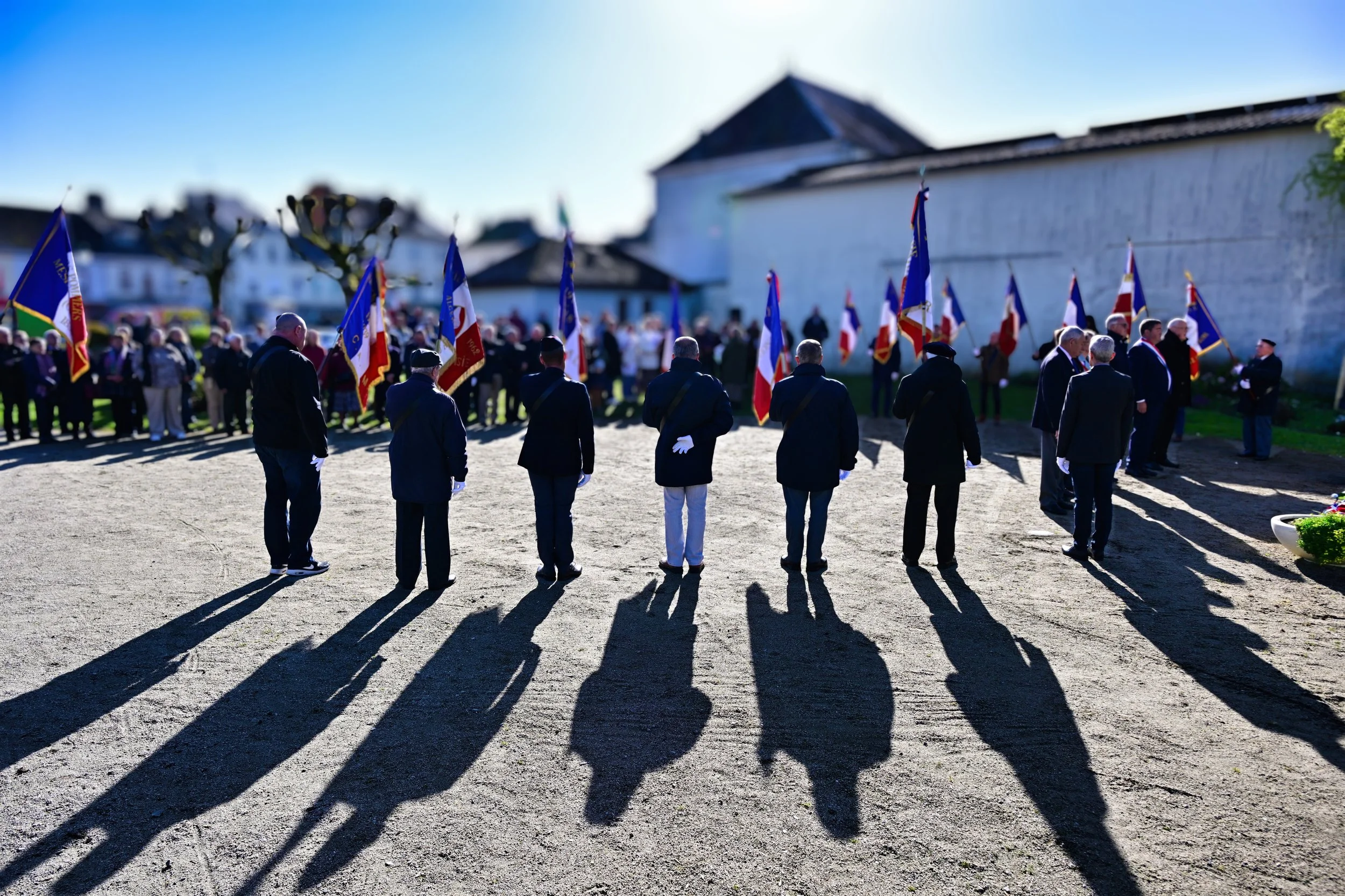 Un groupe de personnes en uniforme assiste à une cérémonie en plein air, certaines portant des drapeaux français, avec de longues ombres projetées par le soleil et un bâtiment en pierre en arrière-plan.