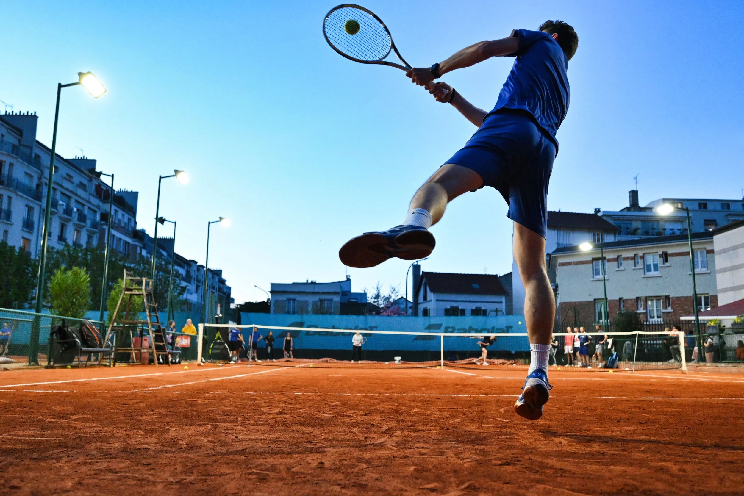Un joueur de tennis en action dans un terrain en extérieur, avec une foule en arrière-plan et des lampadaires illuminant la scène, lors d'une soirée