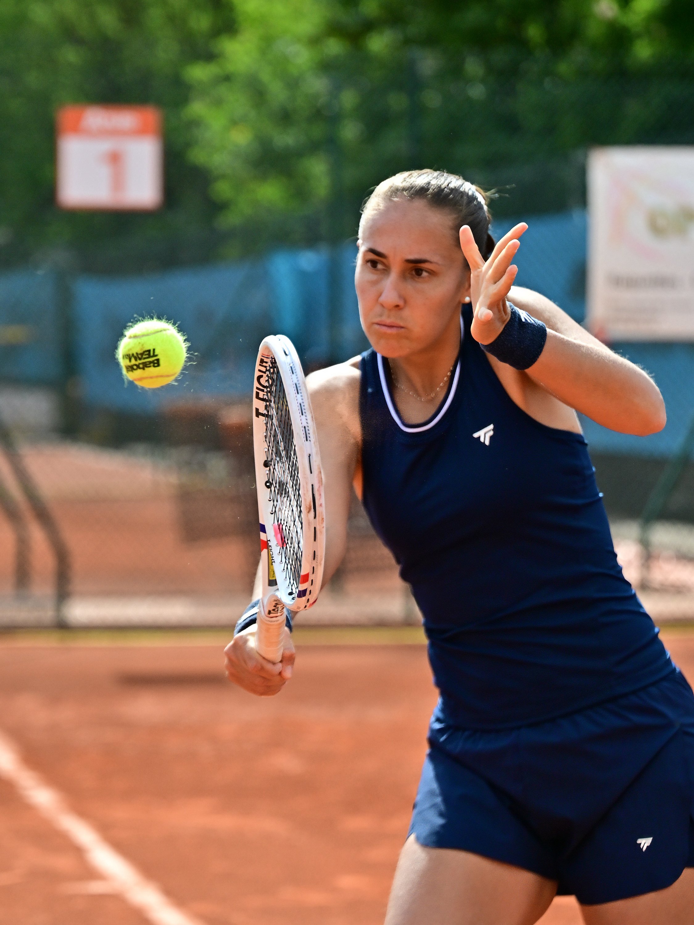 Une femme joue au tennis sur un court en terre battue, frappant une balle jaune avec une raquette.