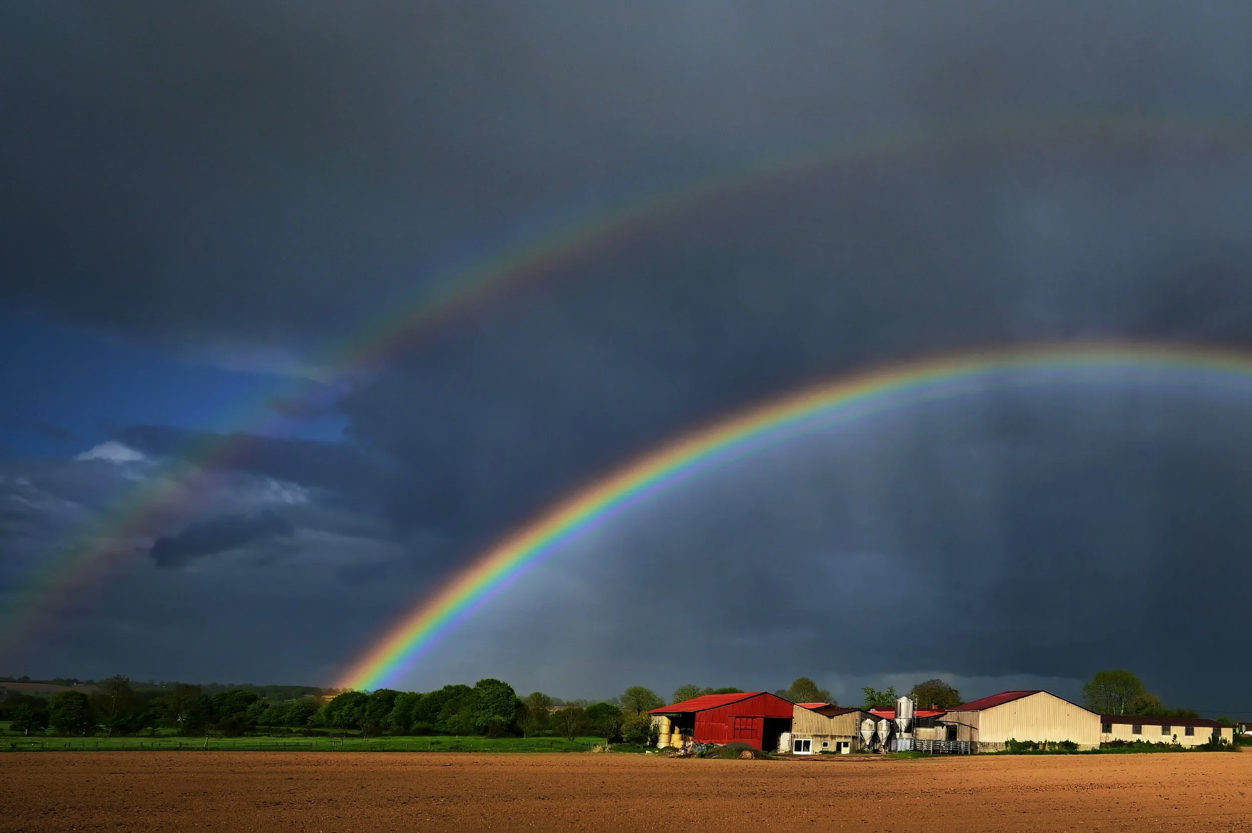 Paysage rural avec trois arcs-en-ciel dans un ciel nuageux, ferme rouge et bâtiments agricoles en arrière-plan.
