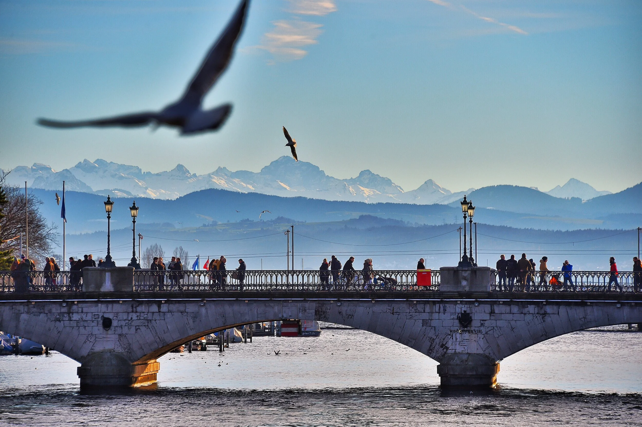 Une scène en bord de rivière avec un pont en pierre, des personnes se promenant, des oiseaux volant dans le ciel, et des montagnes enneigées en arrière-plan.